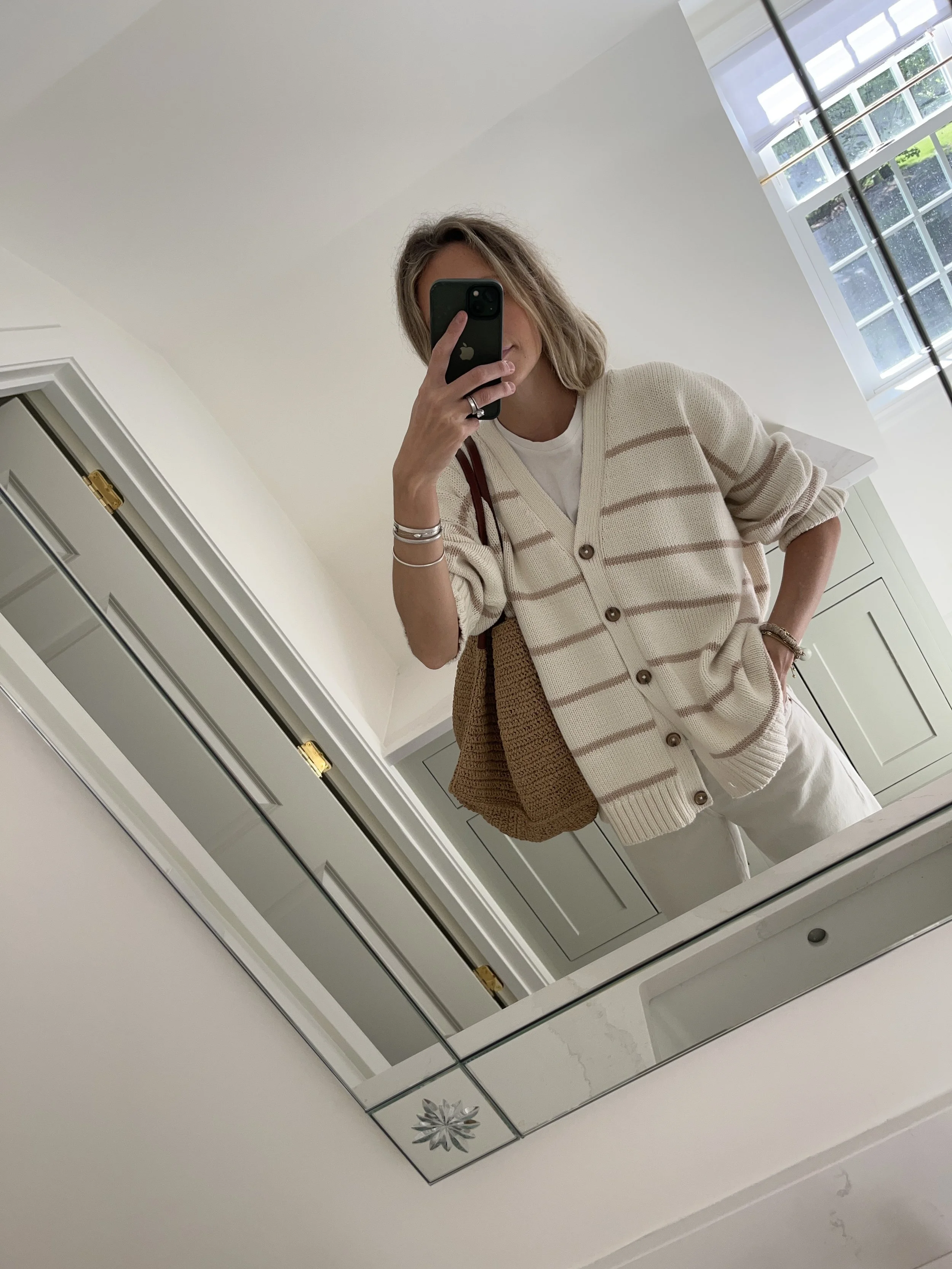 Person taking a mirror selfie in a living room with hardwood floors, wearing a black fleece jacket, gray shirt, black leggings, gray sneakers, and a white cap with a logo.