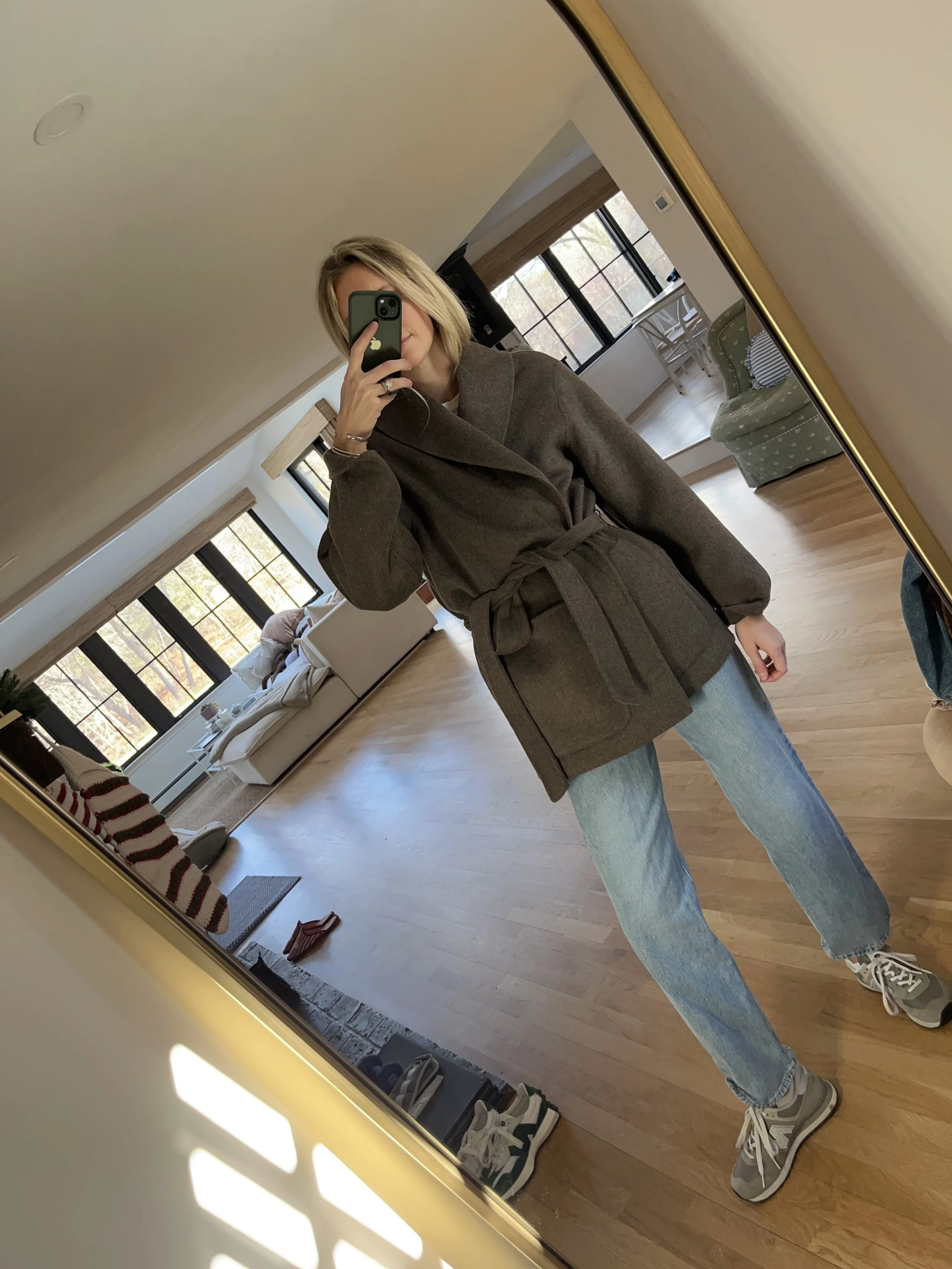 A woman taking a mirror selfie in a spacious living room with wooden floors, large windows, and minimal furniture, wearing a beige jacket, black top, blue jeans, and tan shoes.