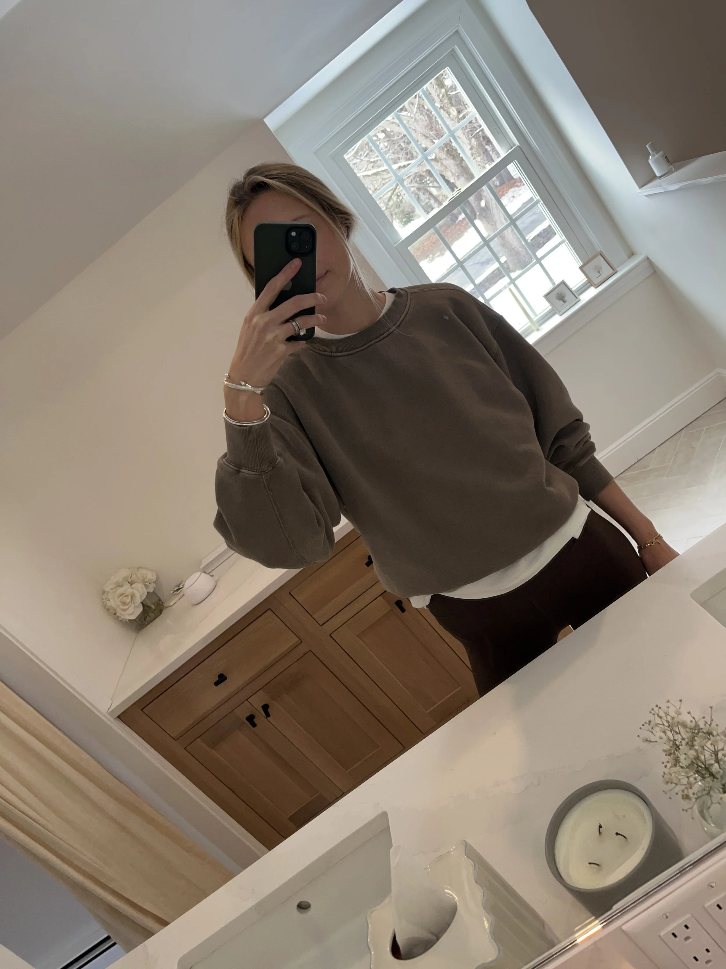 Person taking a mirror selfie in a living room with hardwood floors, wearing a black fleece jacket, gray shirt, black leggings, gray sneakers, and a white cap with a logo.