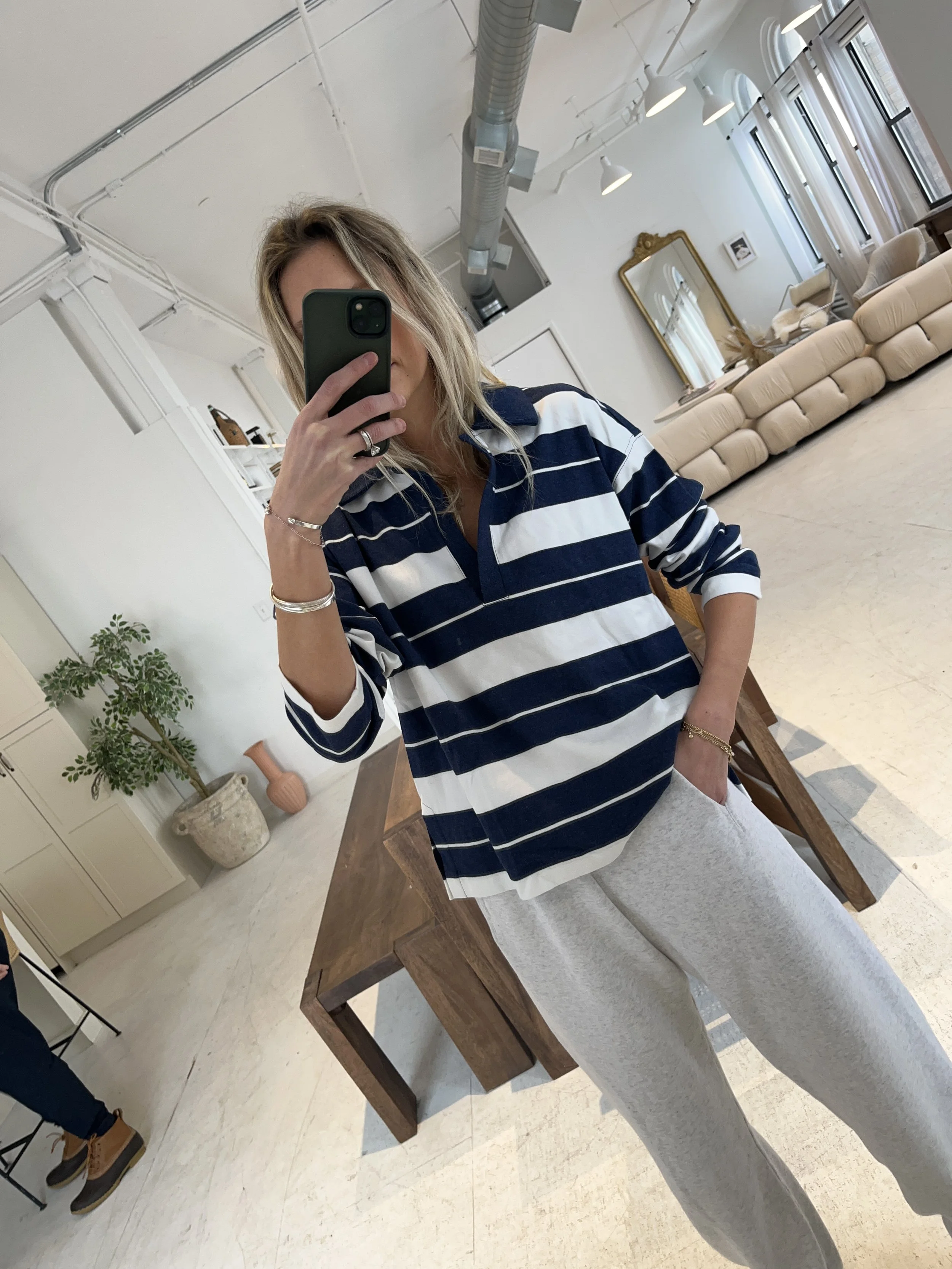 A woman taking a mirror selfie in a spacious, well-lit living room with hardwood floors, large windows, a white sofa, and a side table with a plant.