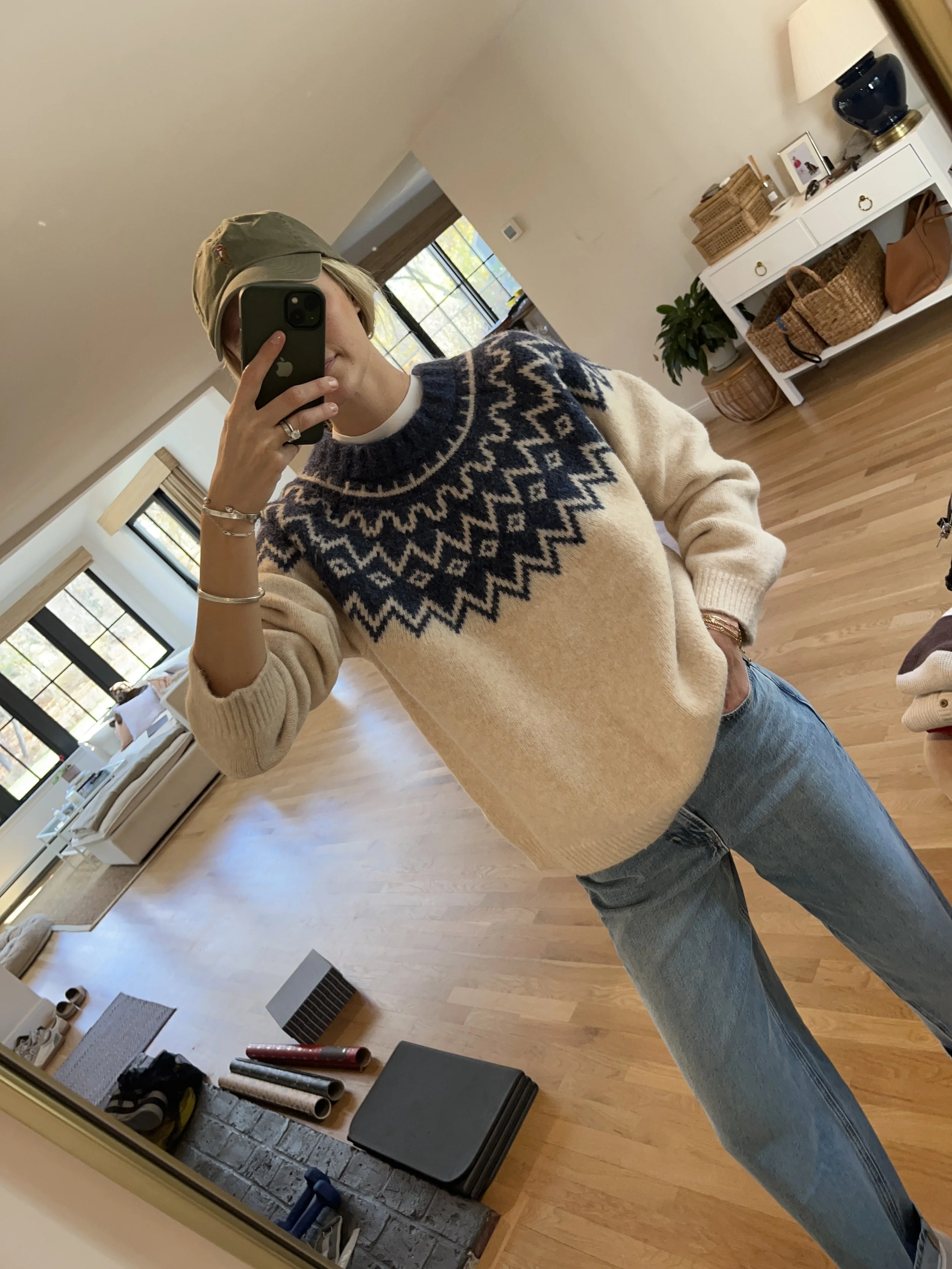 Person taking a mirror selfie in a living room with hardwood floors, wearing a black fleece jacket, gray shirt, black leggings, gray sneakers, and a white cap with a logo.