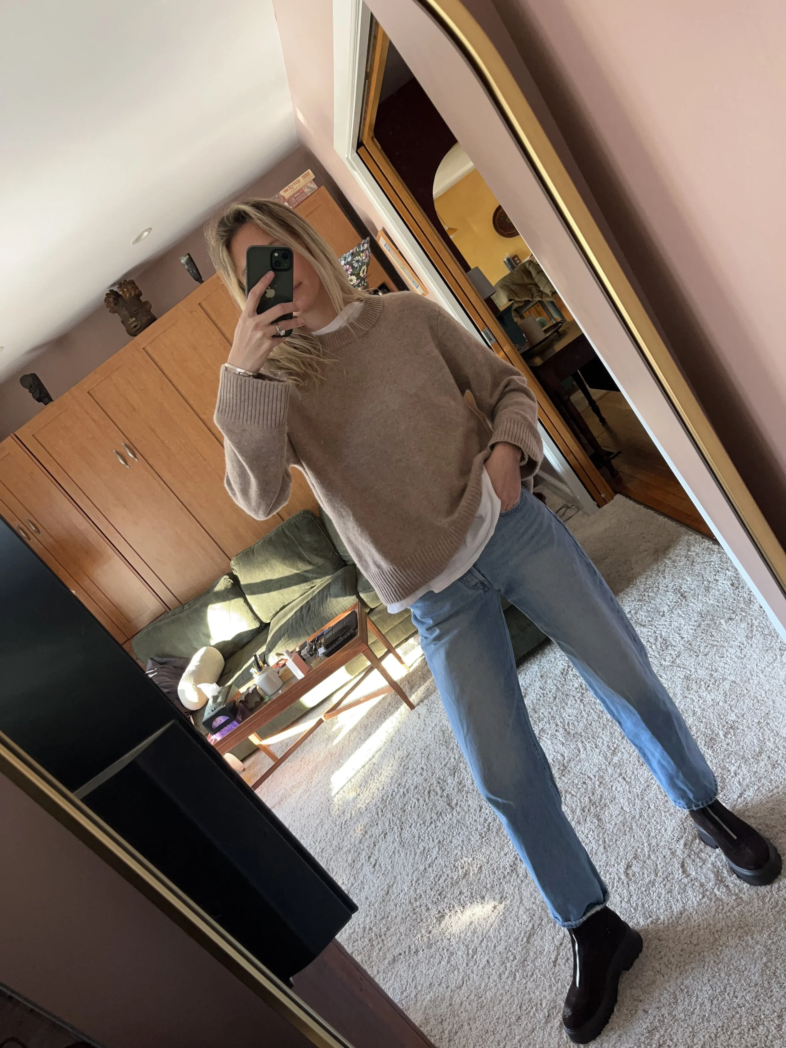 Person taking a mirror selfie in a living room with hardwood floors, wearing a black fleece jacket, gray shirt, black leggings, gray sneakers, and a white cap with a logo.