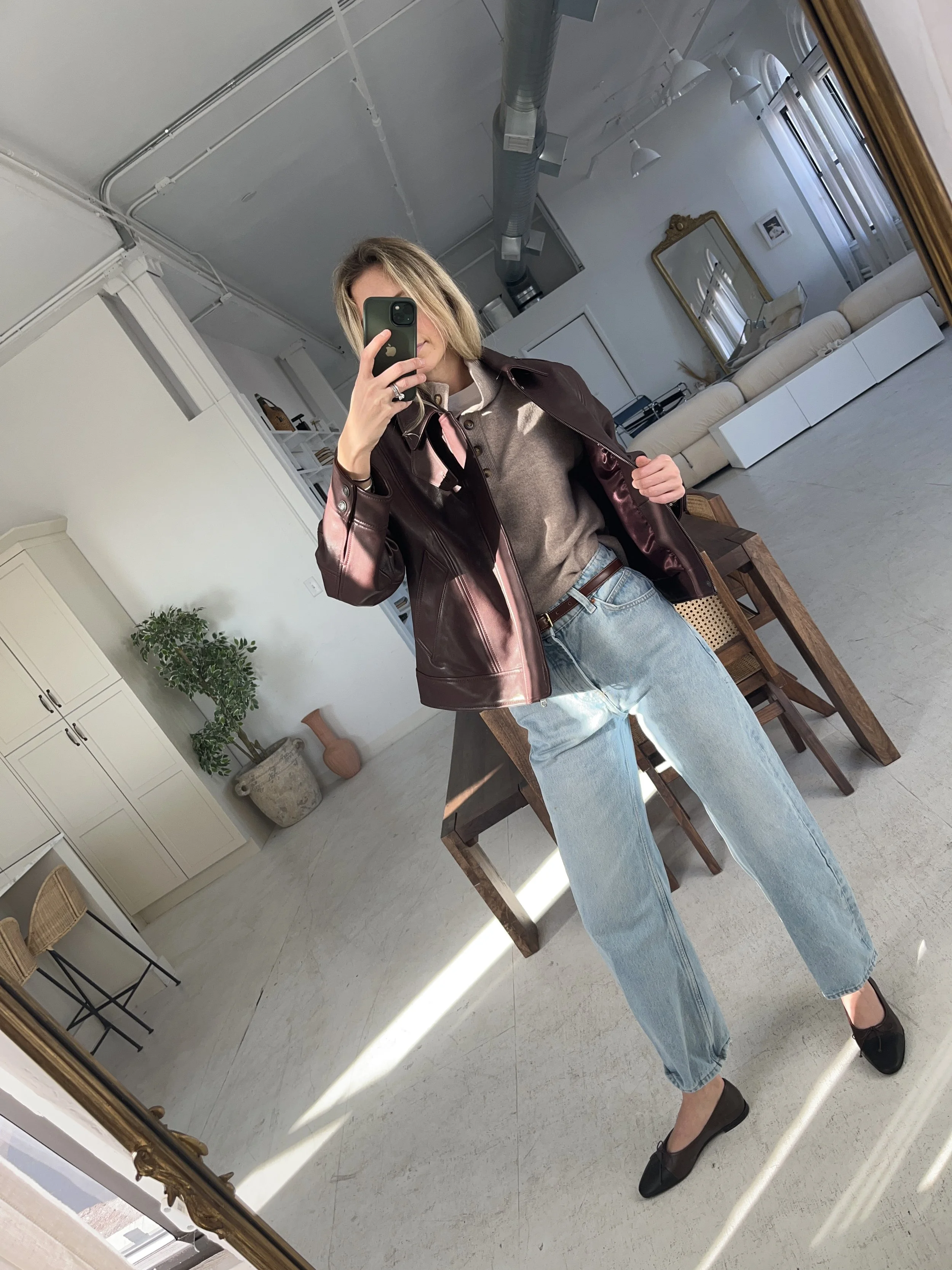 Person taking a mirror selfie in a living room with hardwood floors, wearing a black fleece jacket, gray shirt, black leggings, gray sneakers, and a white cap with a logo.