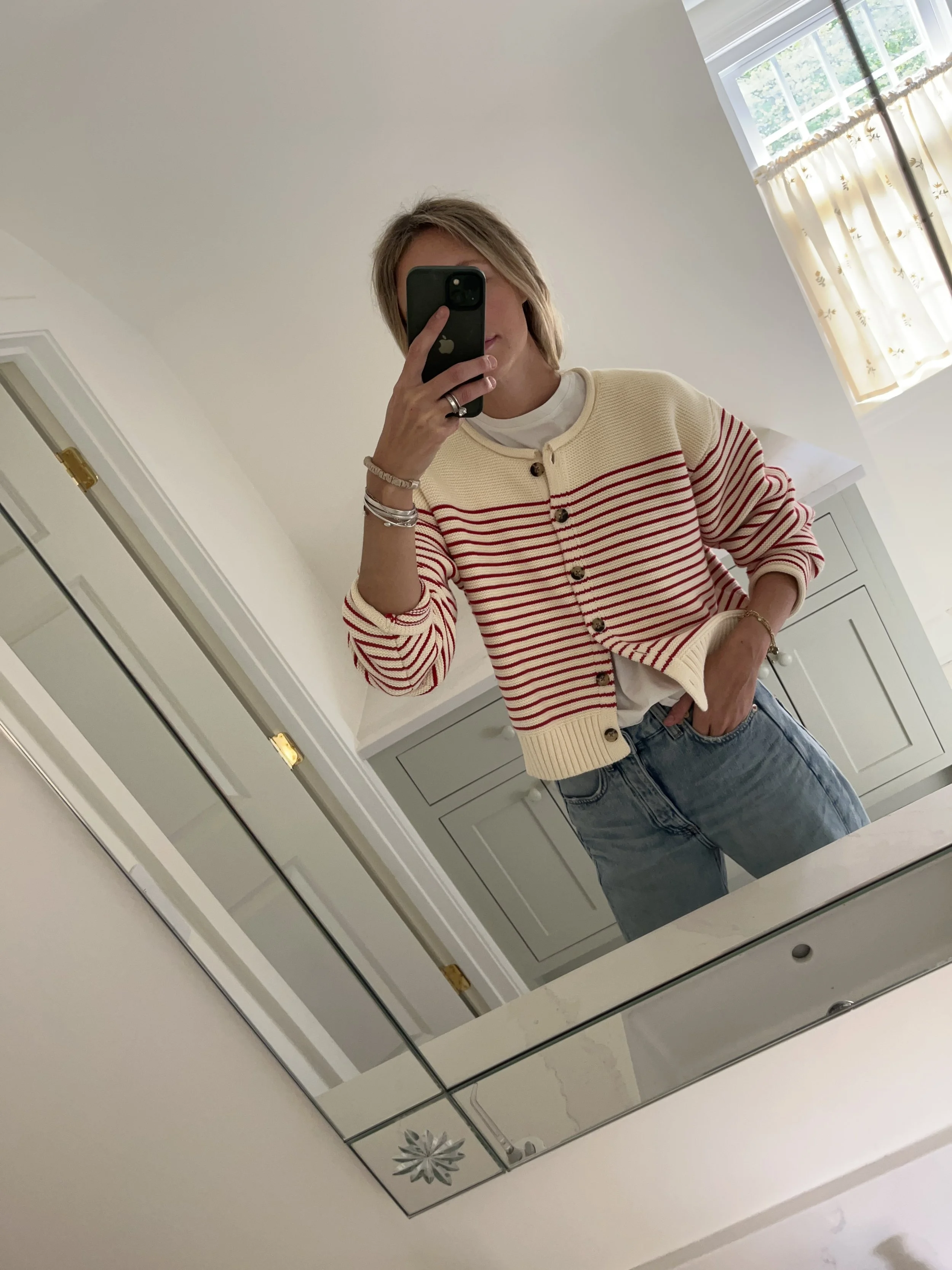 Person taking a mirror selfie in a room with wooden floors, wearing a striped brown and white long-sleeve shirt, light blue jeans, and black and white sneakers.