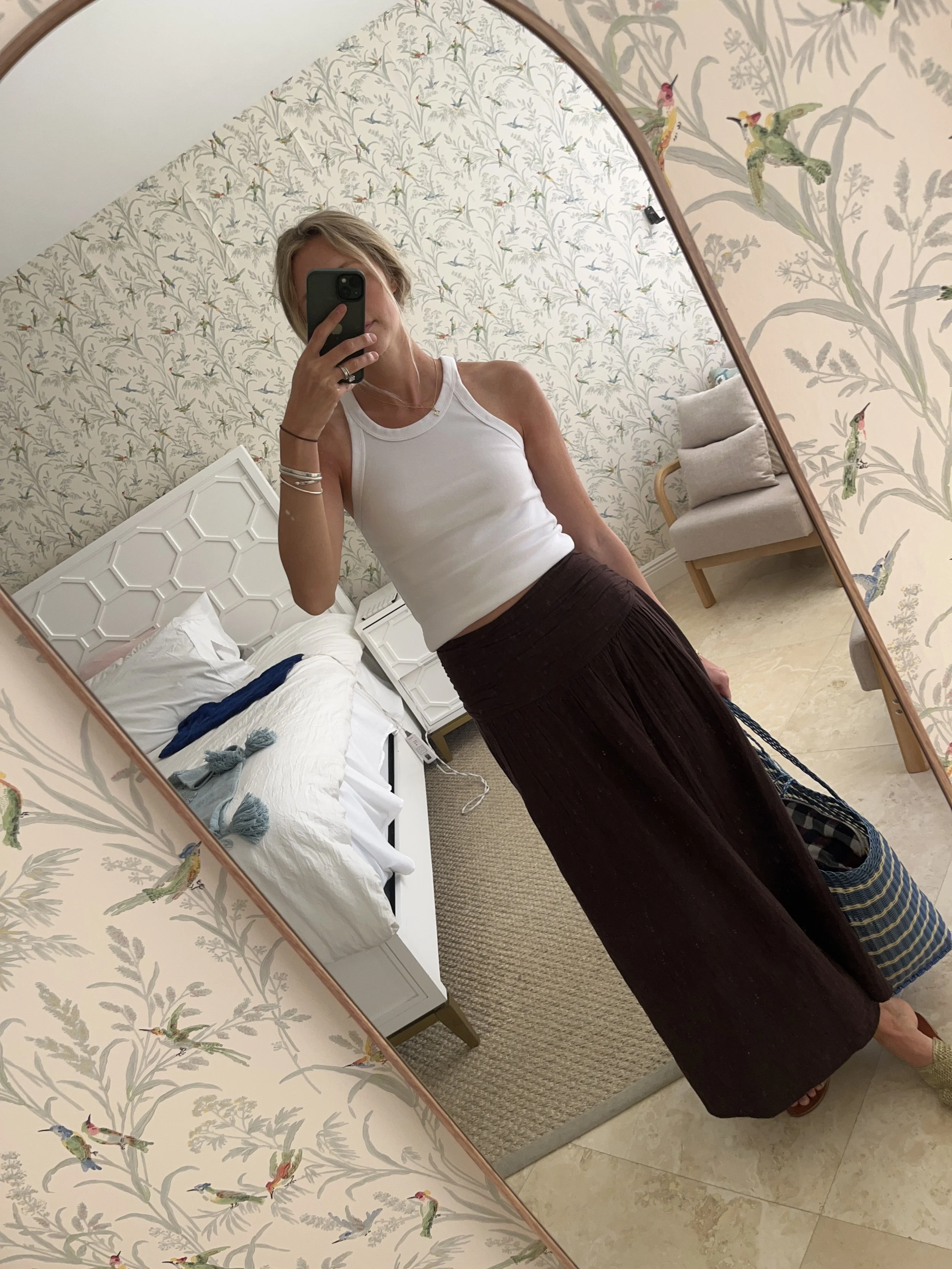 A woman taking a mirror selfie in a spacious, well-lit living room with hardwood floors, large windows, a white sofa, and a side table with a plant.