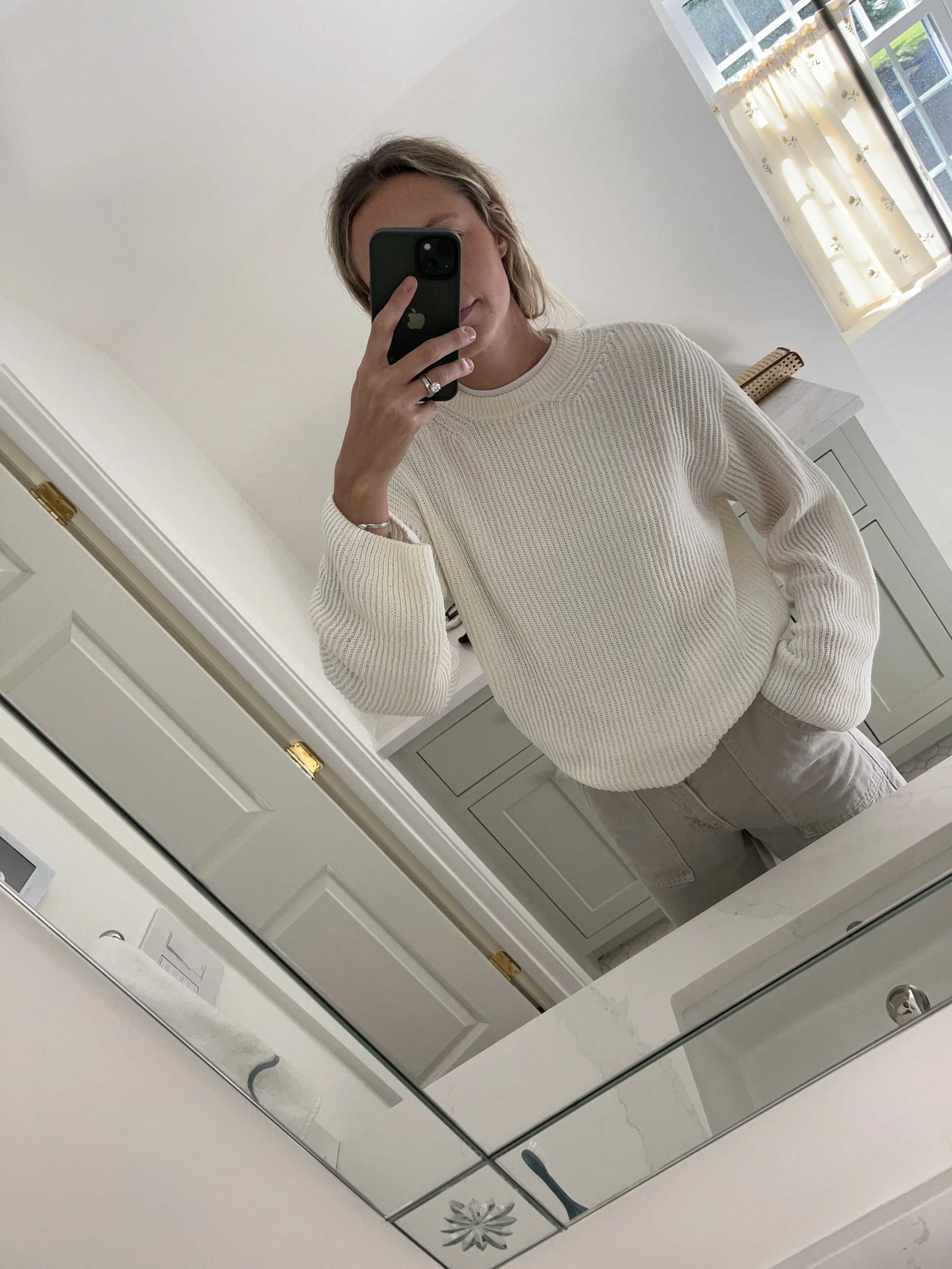 Person taking a mirror selfie in a living room with hardwood floors, wearing a black fleece jacket, gray shirt, black leggings, gray sneakers, and a white cap with a logo.