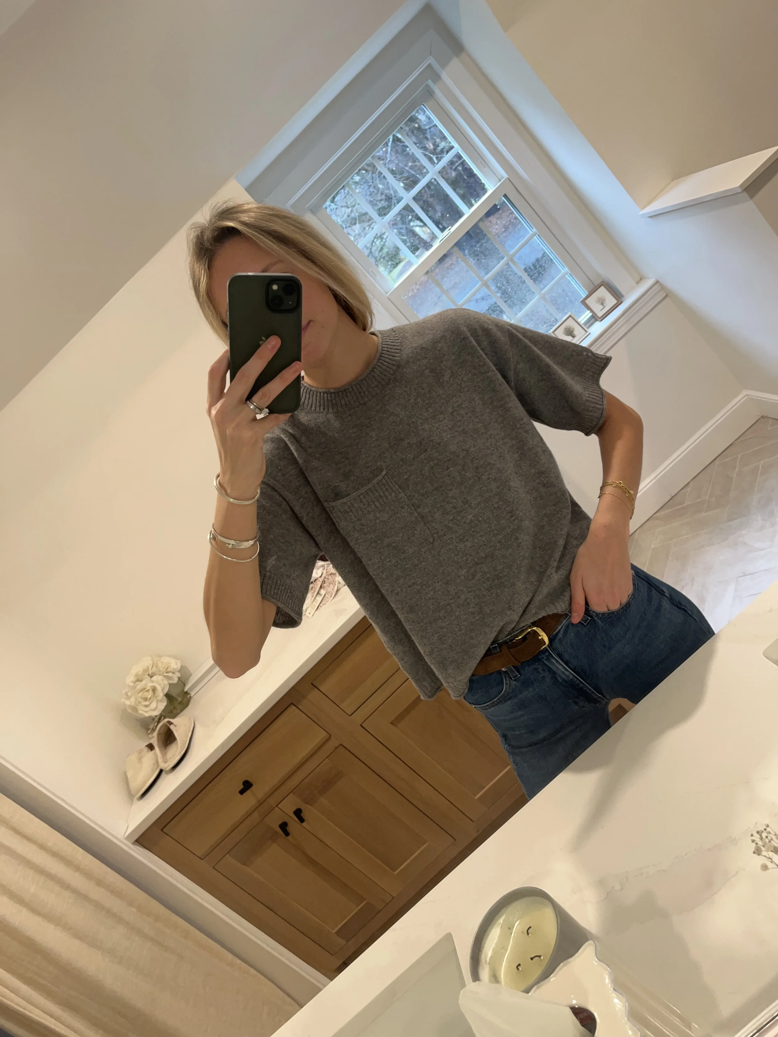 A woman taking a mirror selfie in a spacious living room with wooden floors, large windows, and minimal furniture, wearing a beige jacket, black top, blue jeans, and tan shoes.