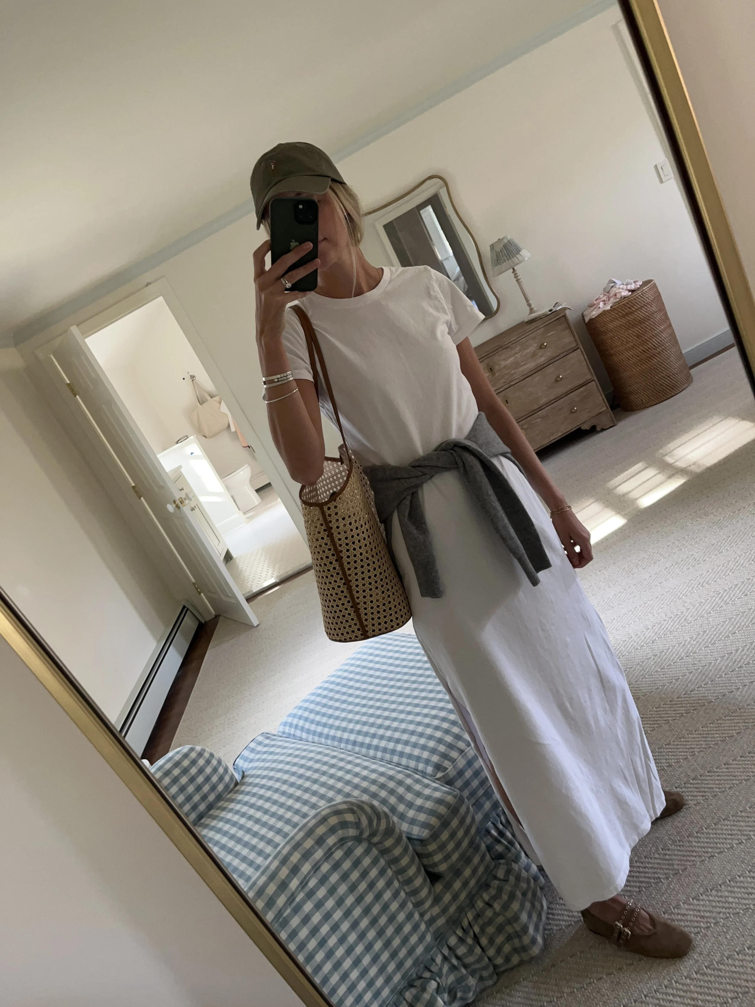 A woman taking a mirror selfie in a spacious, well-lit living room with hardwood floors, large windows, a white sofa, and a side table with a plant.