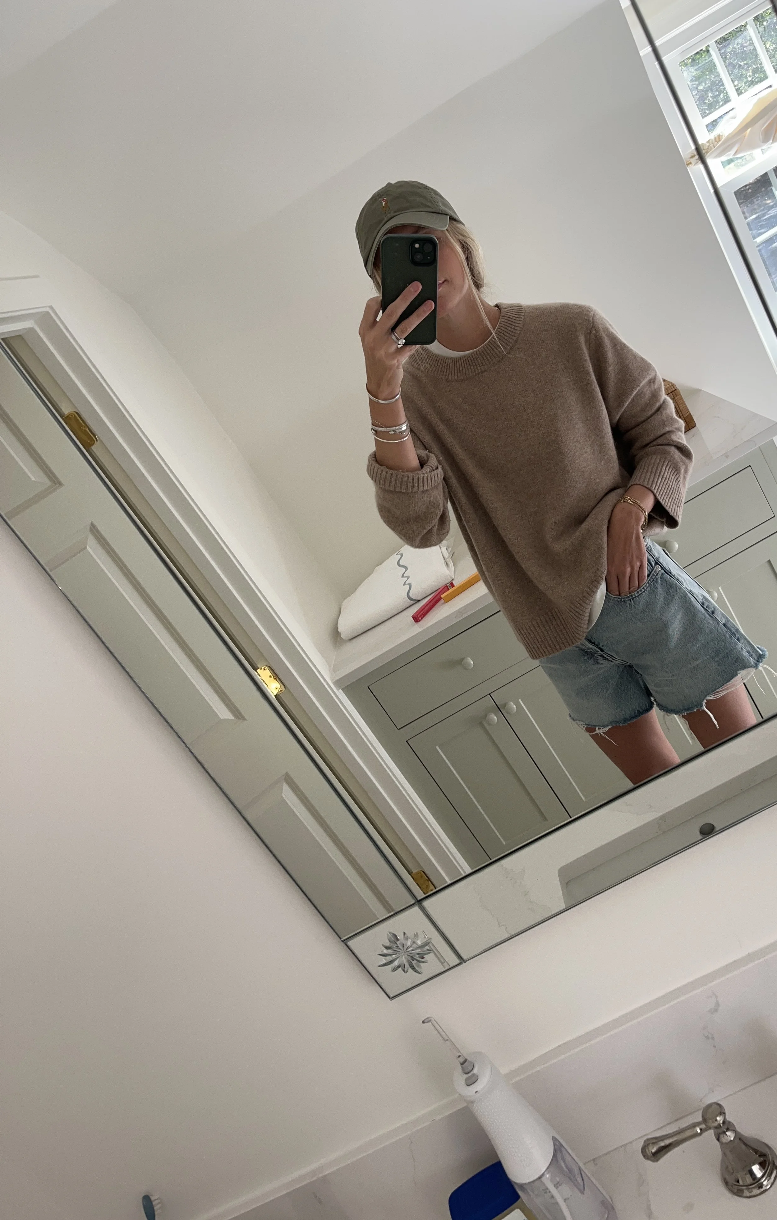A woman taking a mirror selfie in a spacious living room with wooden floors, large windows, and minimal furniture, wearing a beige jacket, black top, blue jeans, and tan shoes.