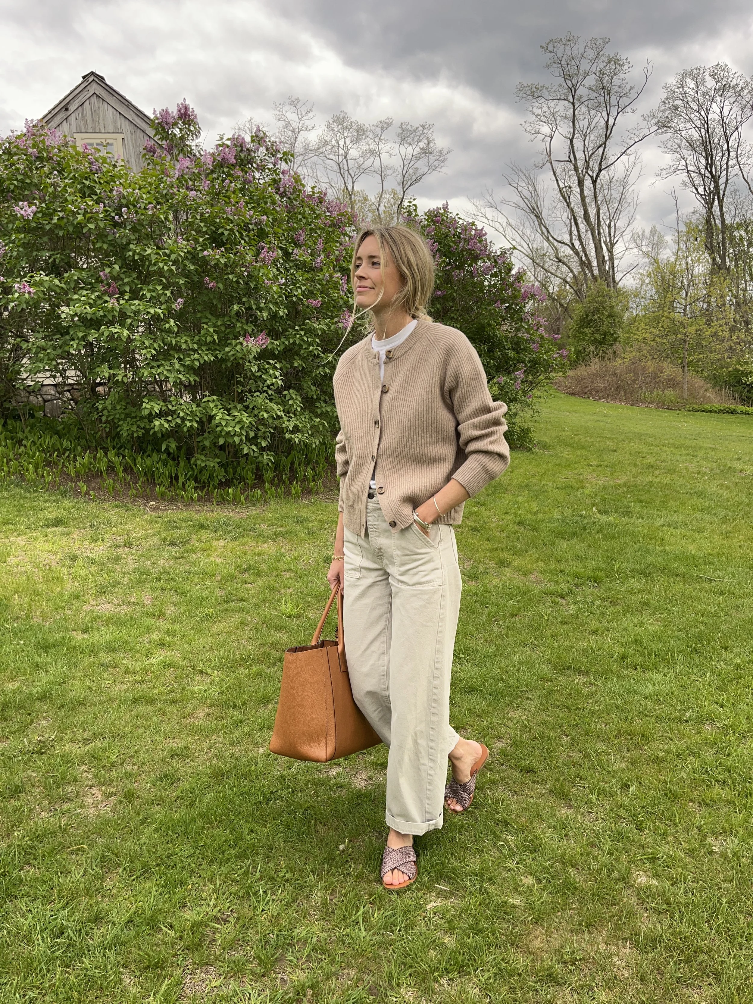 A woman taking a mirror selfie in a spacious living room with wooden floors, large windows, and minimal furniture, wearing a beige jacket, black top, blue jeans, and tan shoes.