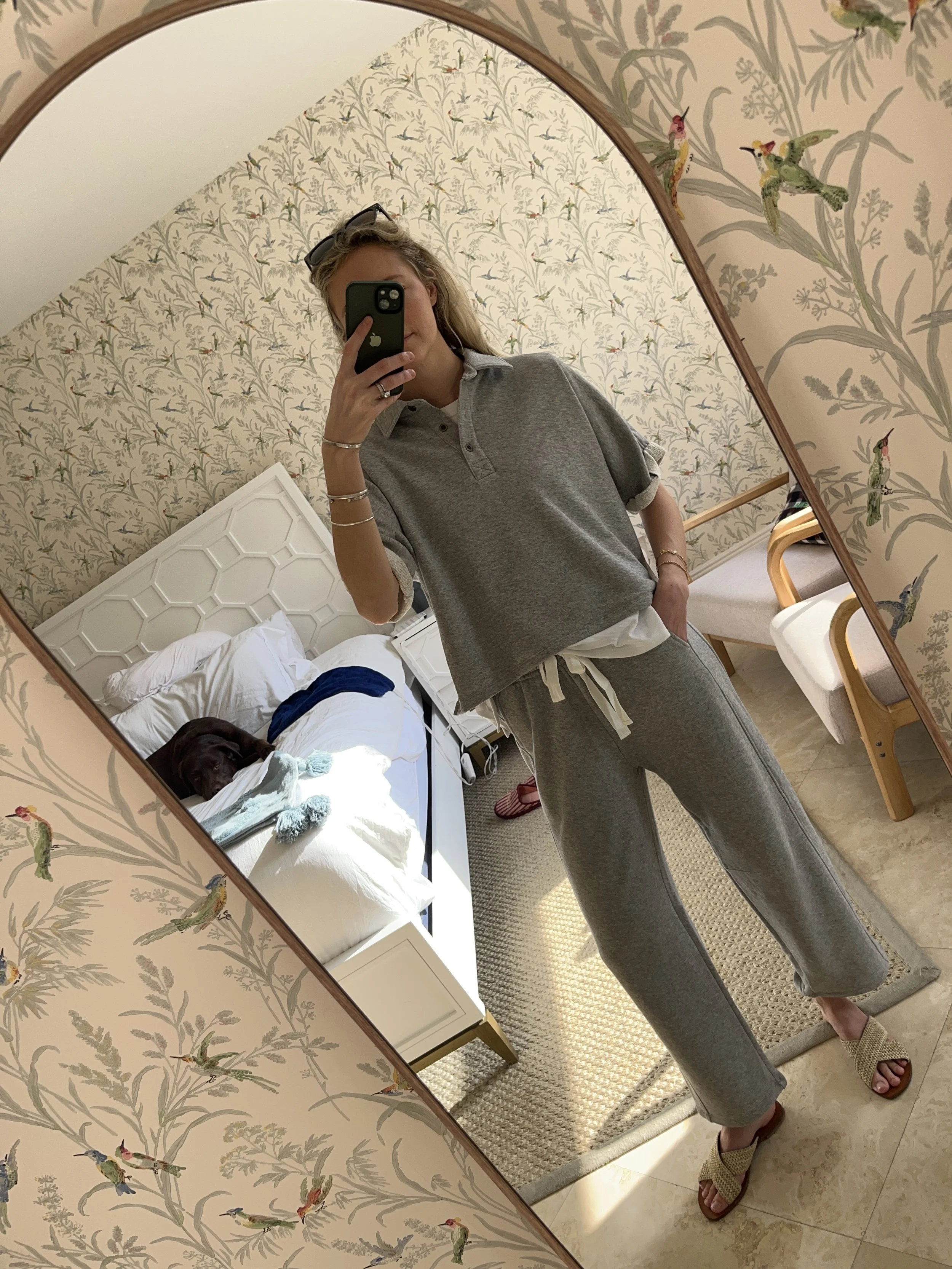 Person taking a mirror selfie in a living room with hardwood floors, wearing a black fleece jacket, gray shirt, black leggings, gray sneakers, and a white cap with a logo.