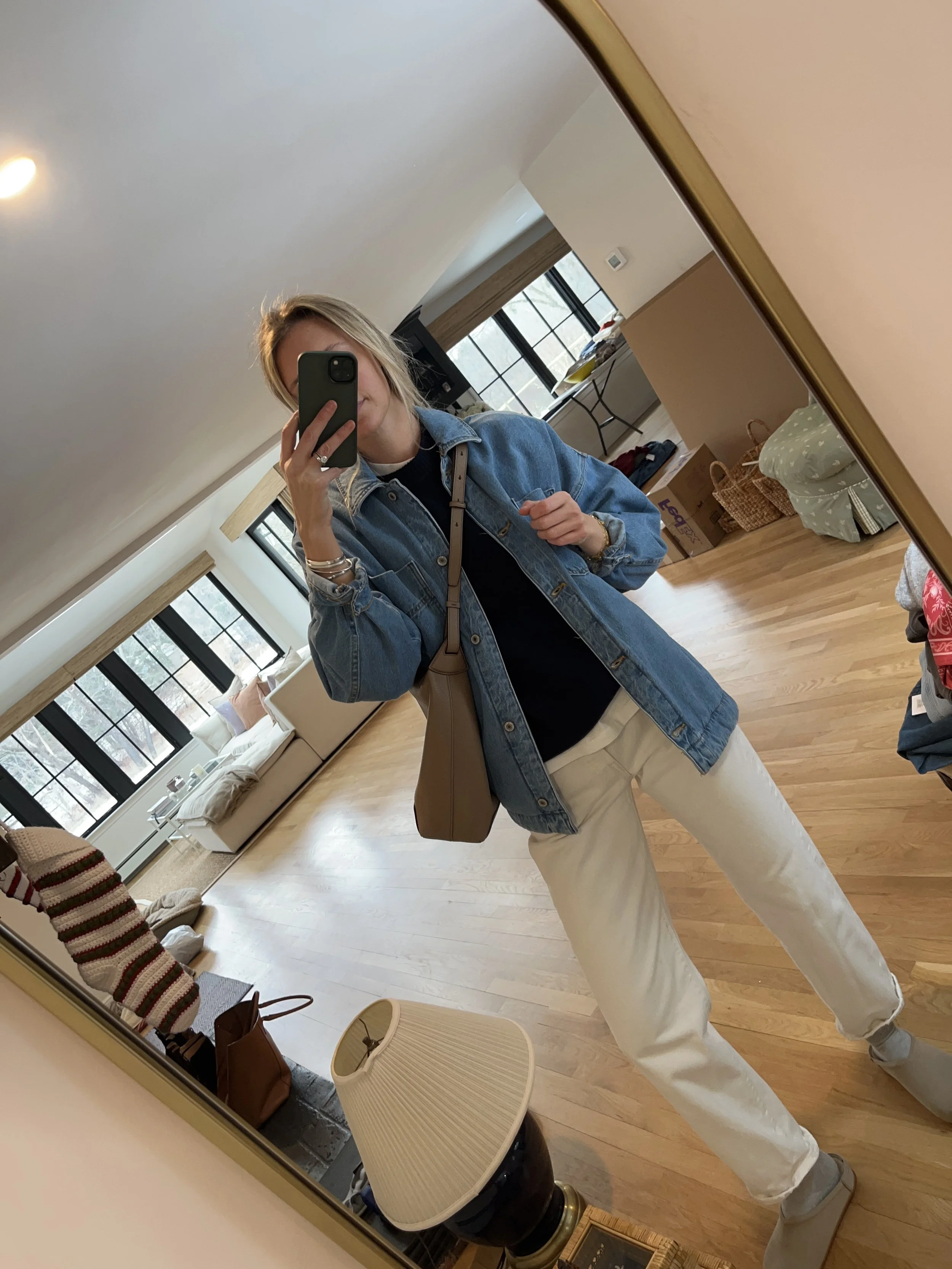 Person taking a mirror selfie in a living room with hardwood floors, wearing a black fleece jacket, gray shirt, black leggings, gray sneakers, and a white cap with a logo.