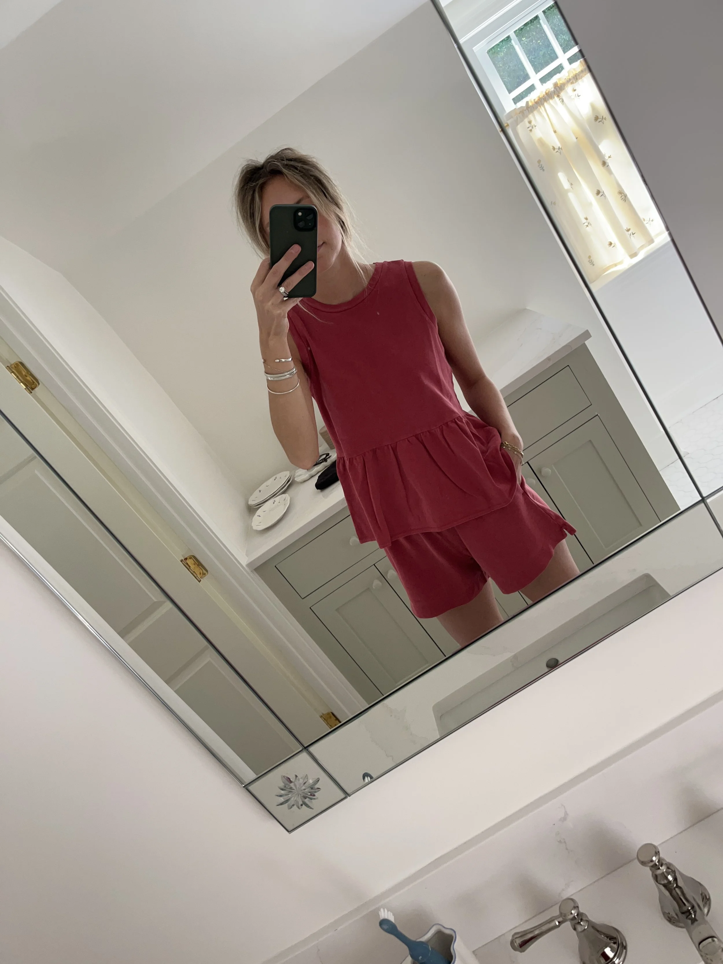 A woman taking a mirror selfie in a spacious living room with wooden floors, large windows, and minimal furniture, wearing a beige jacket, black top, blue jeans, and tan shoes.