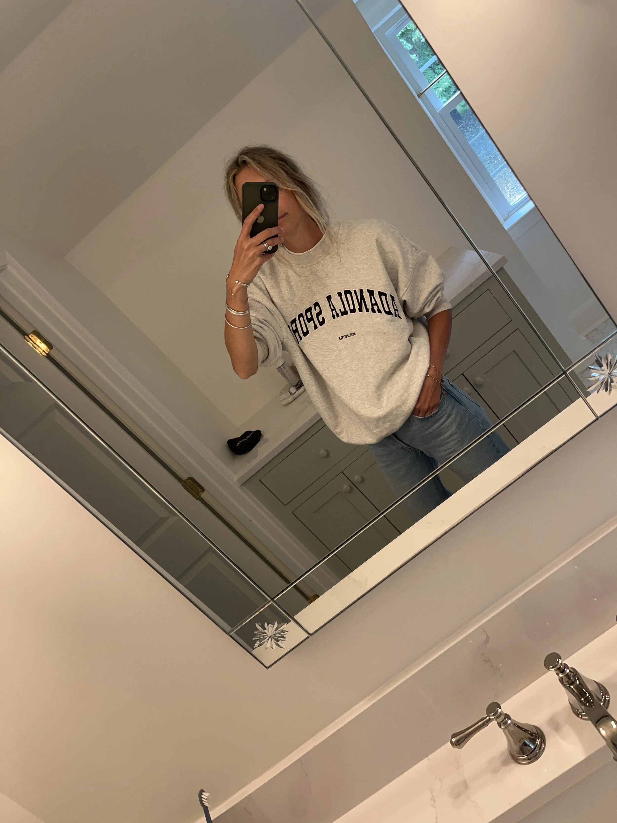 Person taking a mirror selfie in a room with wooden floors, wearing a striped brown and white long-sleeve shirt, light blue jeans, and black and white sneakers.