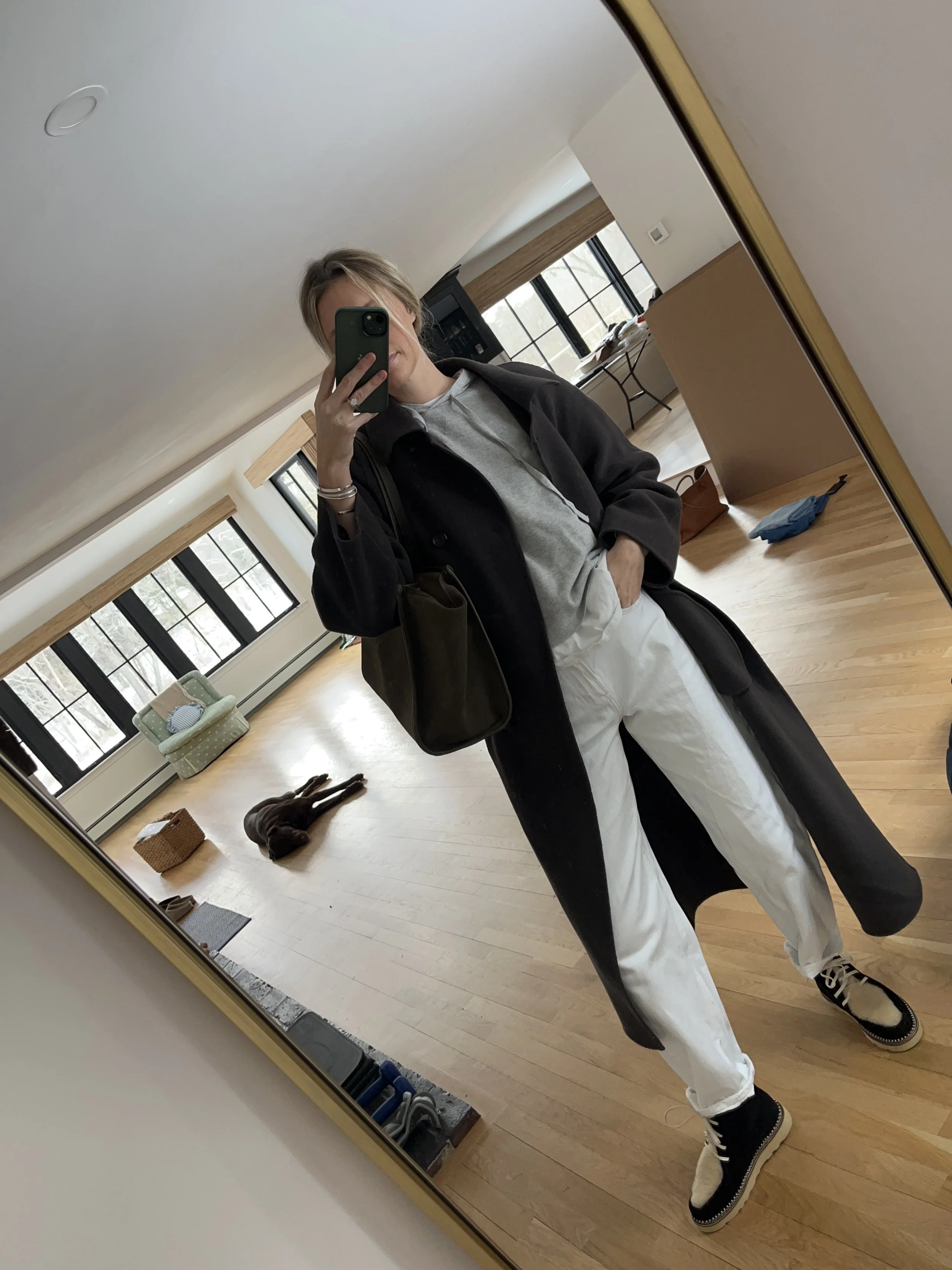 A woman taking a mirror selfie in a spacious living room with wooden floors, large windows, and minimal furniture, wearing a beige jacket, black top, blue jeans, and tan shoes.