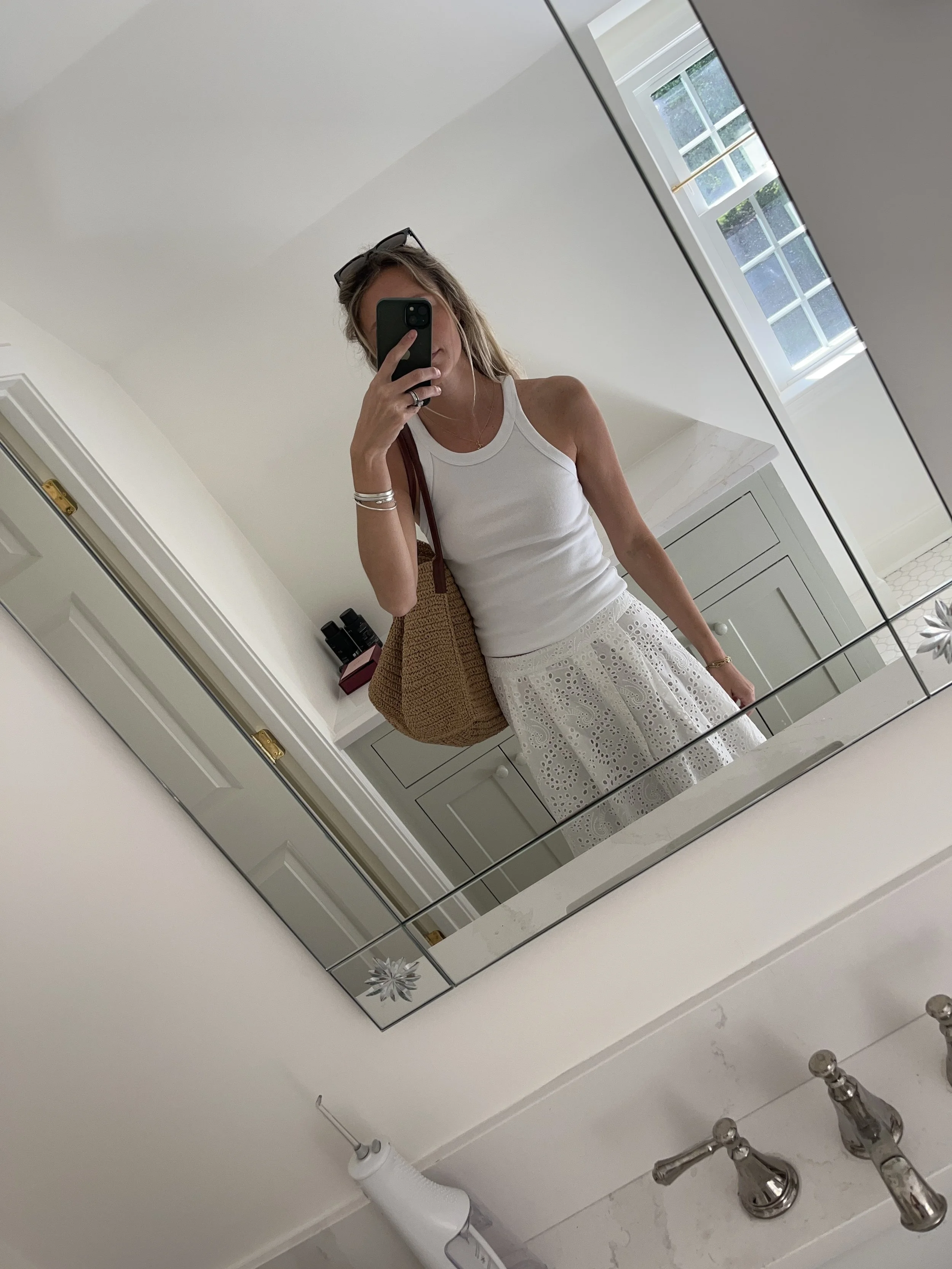 A woman taking a mirror selfie in a spacious, well-lit living room with hardwood floors, large windows, a white sofa, and a side table with a plant.