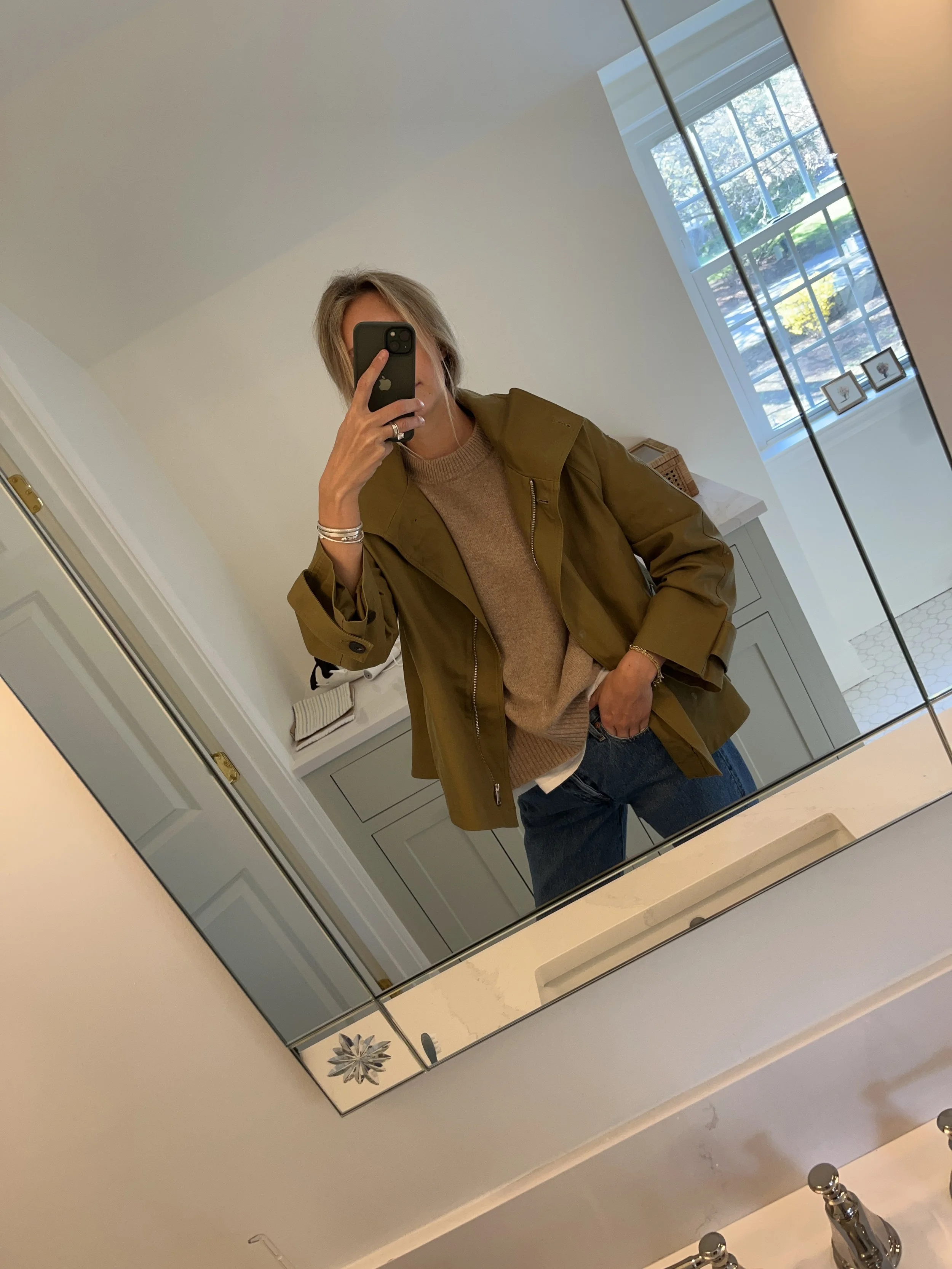 A woman taking a mirror selfie in a spacious living room with wooden floors, large windows, and minimal furniture, wearing a beige jacket, black top, blue jeans, and tan shoes.