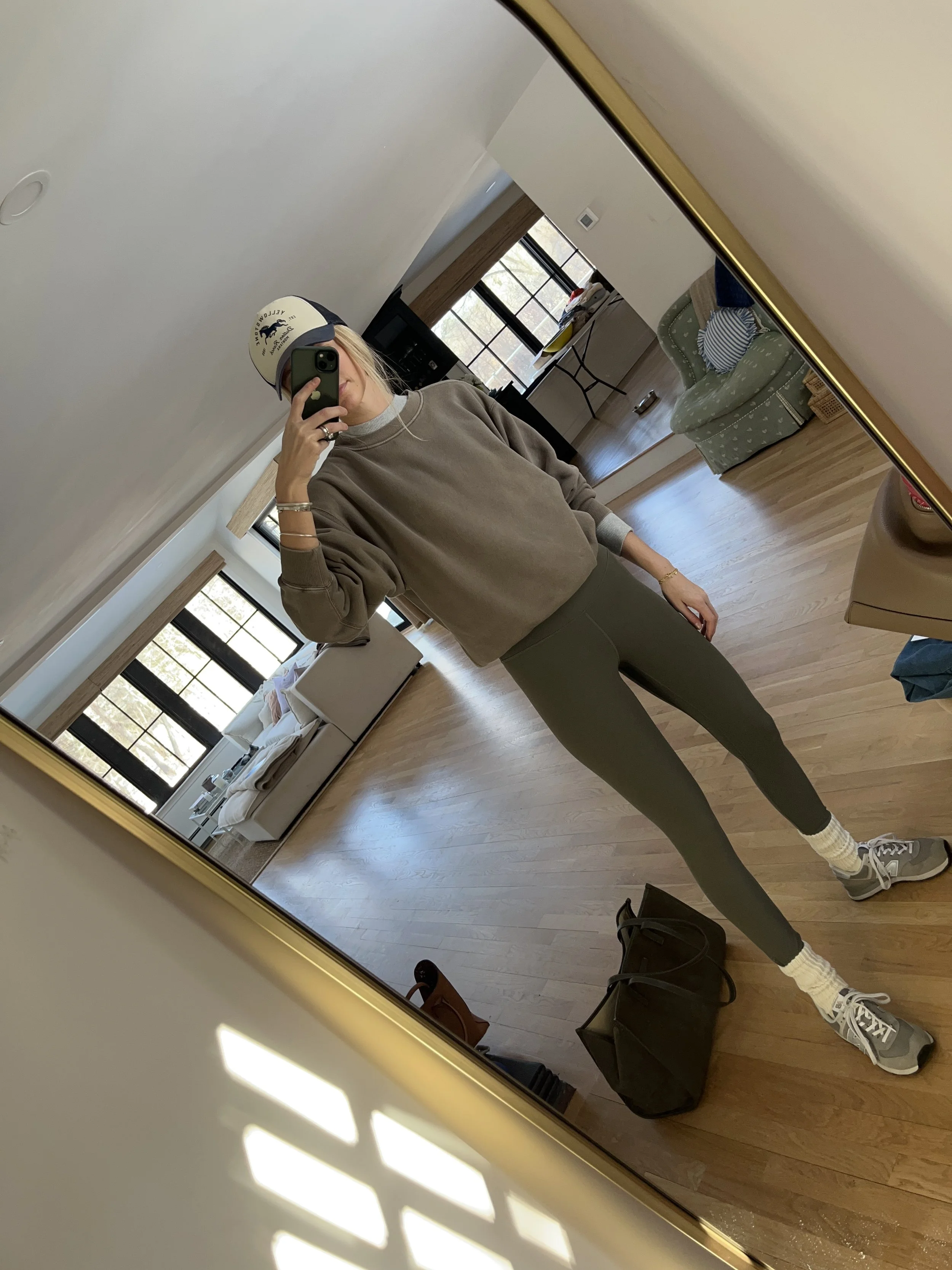 A woman taking a mirror selfie in a spacious living room with wooden floors, large windows, and minimal furniture, wearing a beige jacket, black top, blue jeans, and tan shoes.