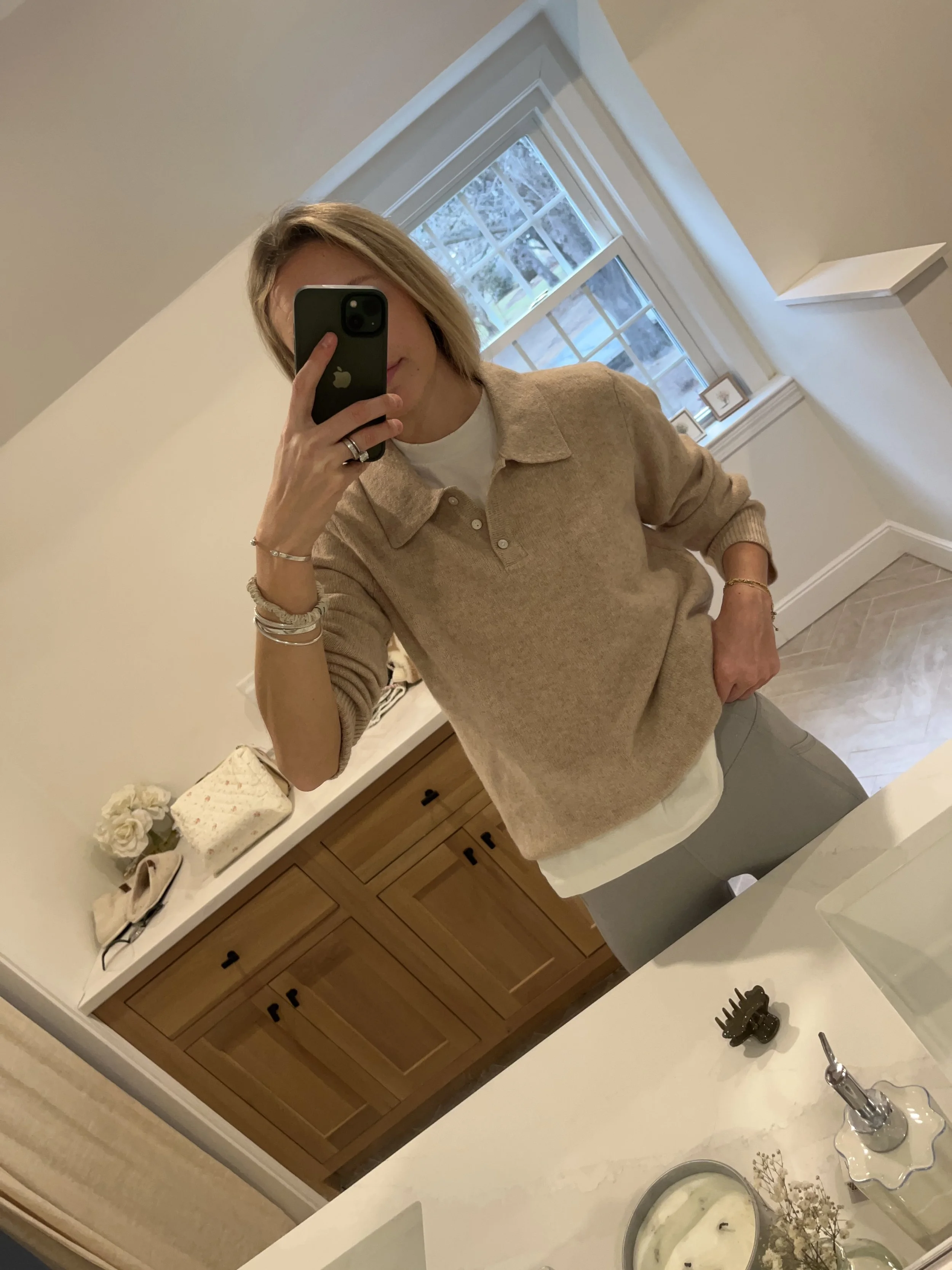 A woman taking a mirror selfie in a spacious, well-lit living room with hardwood floors, large windows, a white sofa, and a side table with a plant.