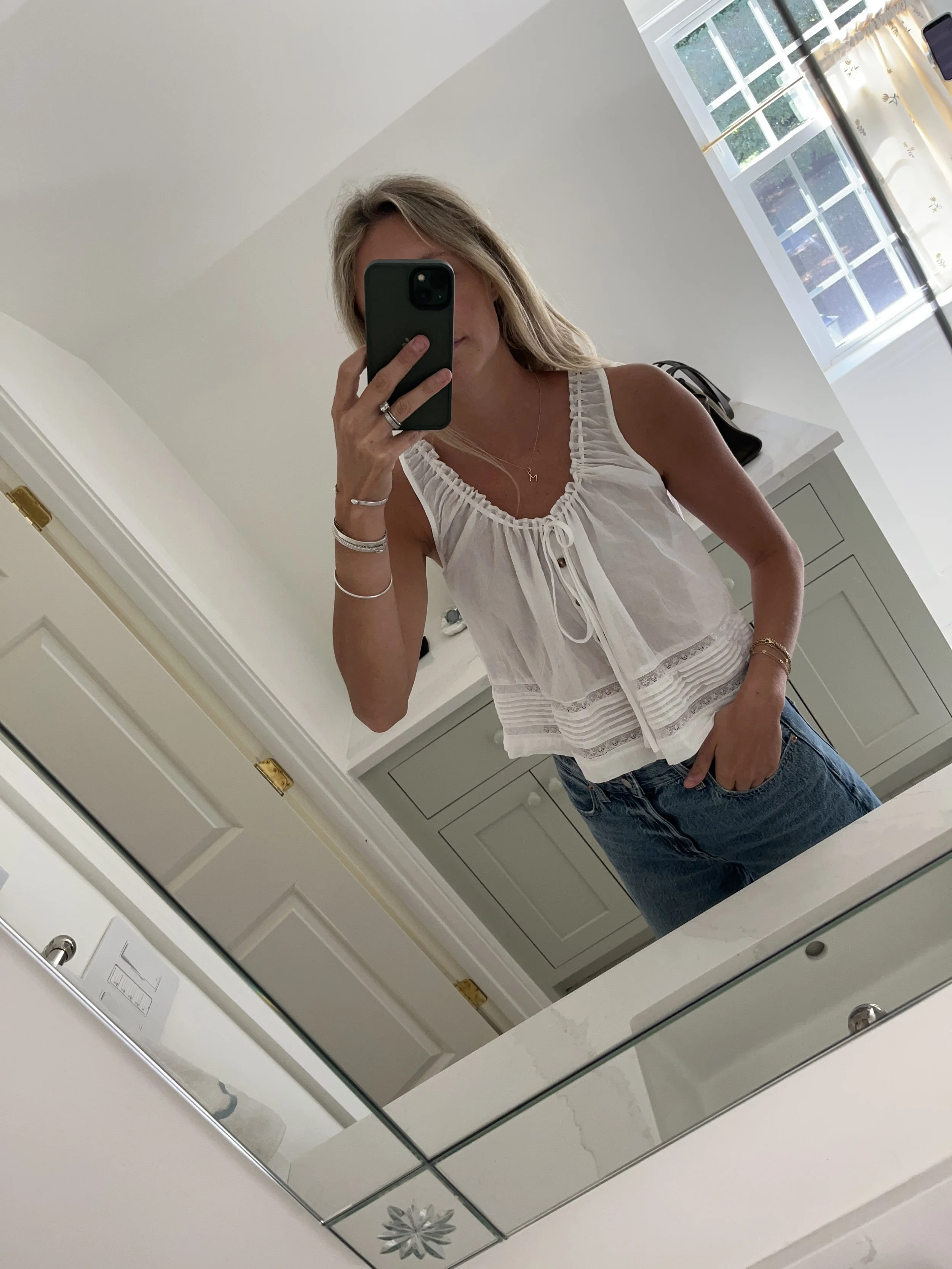 A woman taking a mirror selfie in a spacious living room with wooden floors, large windows, and minimal furniture, wearing a beige jacket, black top, blue jeans, and tan shoes.