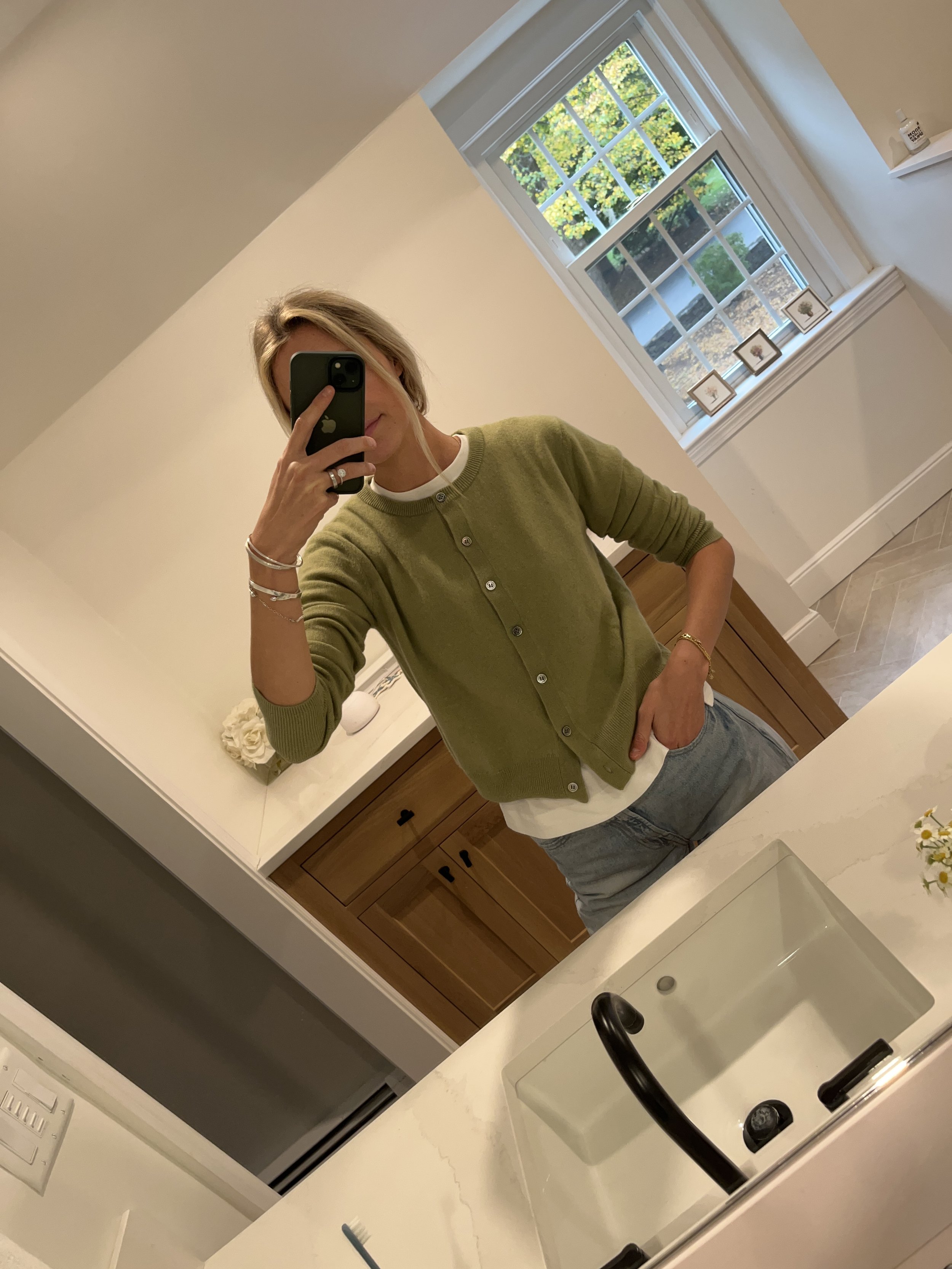 A woman taking a mirror selfie in a bathroom, wearing a green cardigan, white shirt, and gray jeans, with a window showing trees in the background.