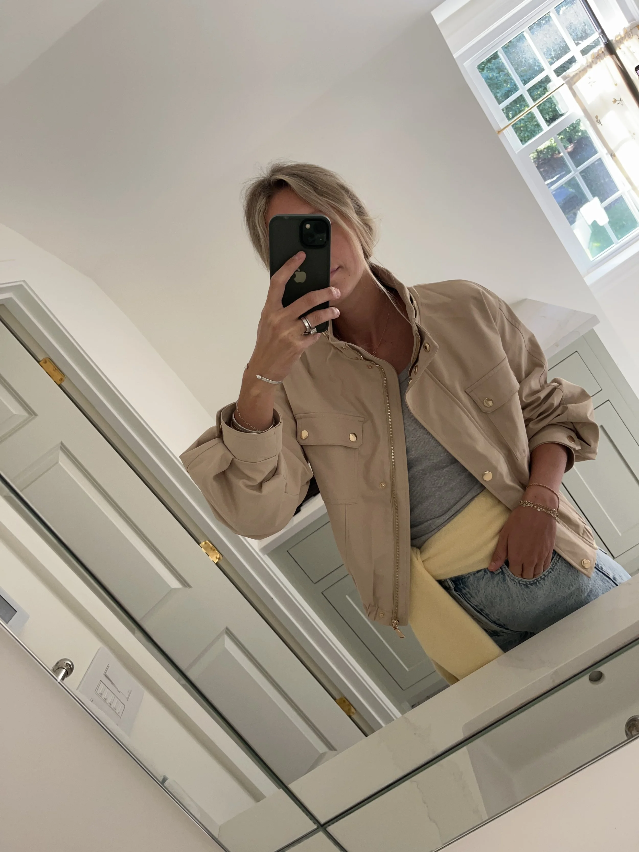 A woman taking a mirror selfie in a spacious, well-lit living room with hardwood floors, large windows, a white sofa, and a side table with a plant.