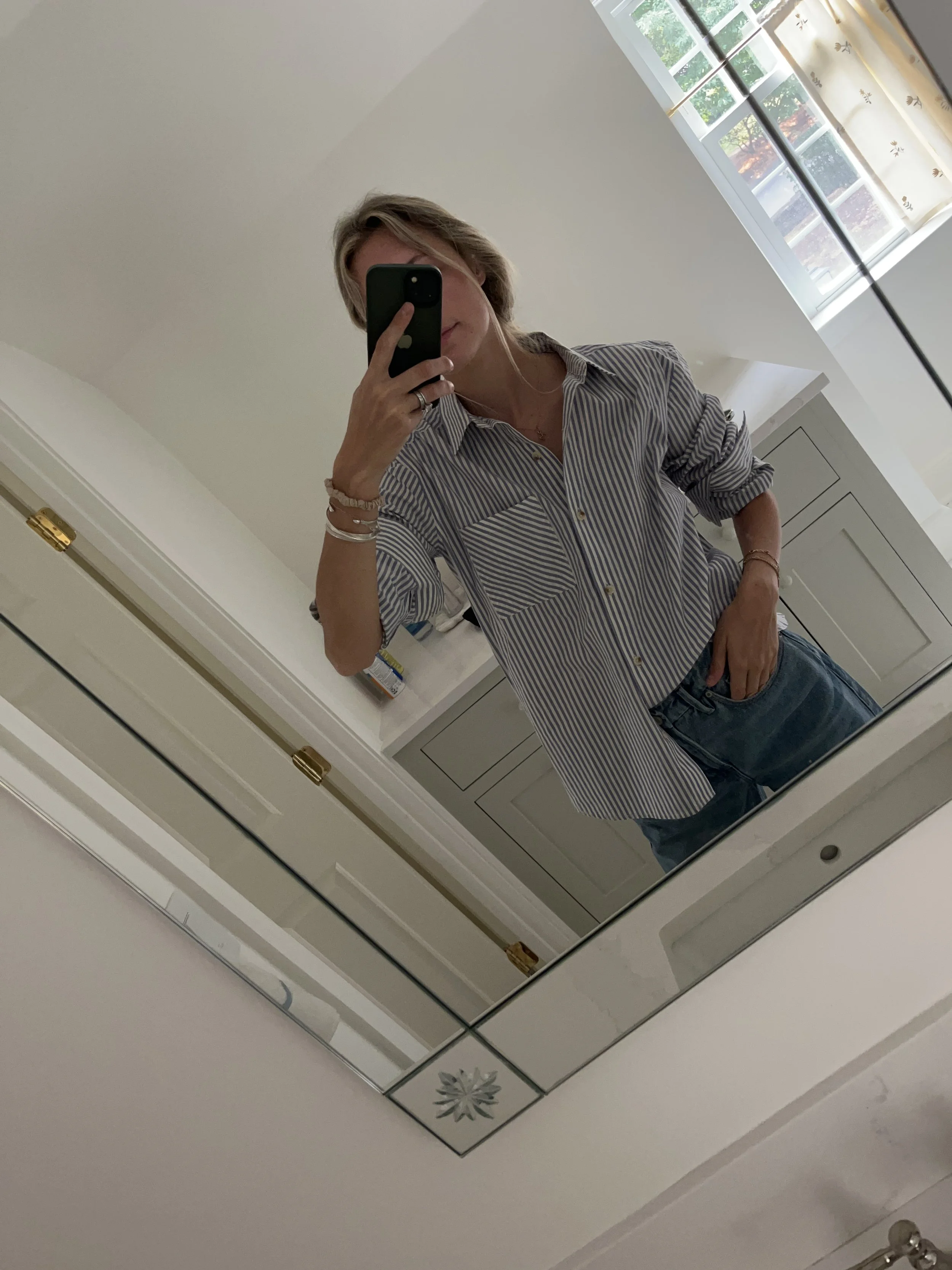 A woman taking a mirror selfie in a spacious living room with wooden floors, large windows, and minimal furniture, wearing a beige jacket, black top, blue jeans, and tan shoes.