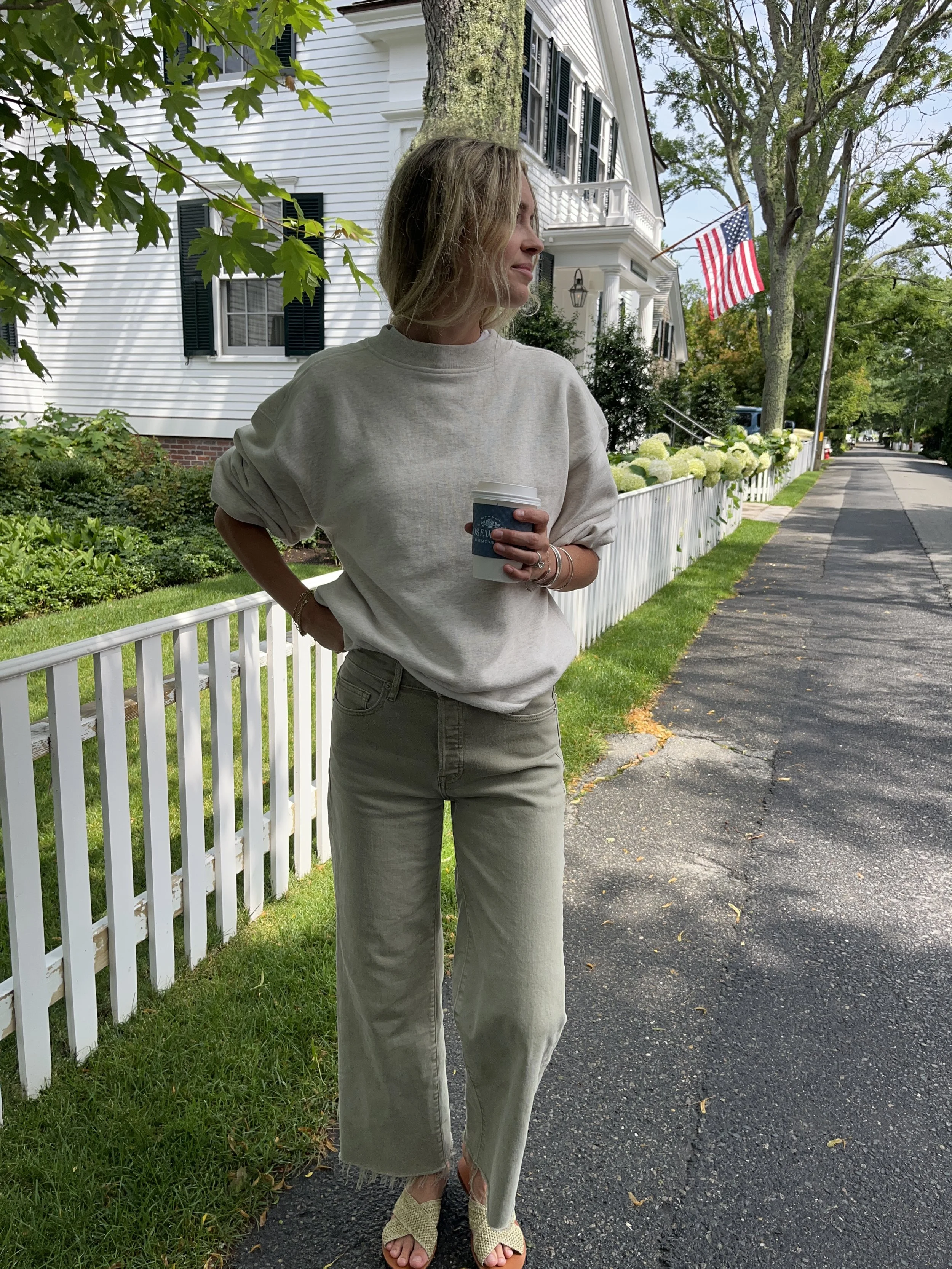 A woman taking a mirror selfie in a spacious living room with wooden floors, large windows, and minimal furniture, wearing a beige jacket, black top, blue jeans, and tan shoes.