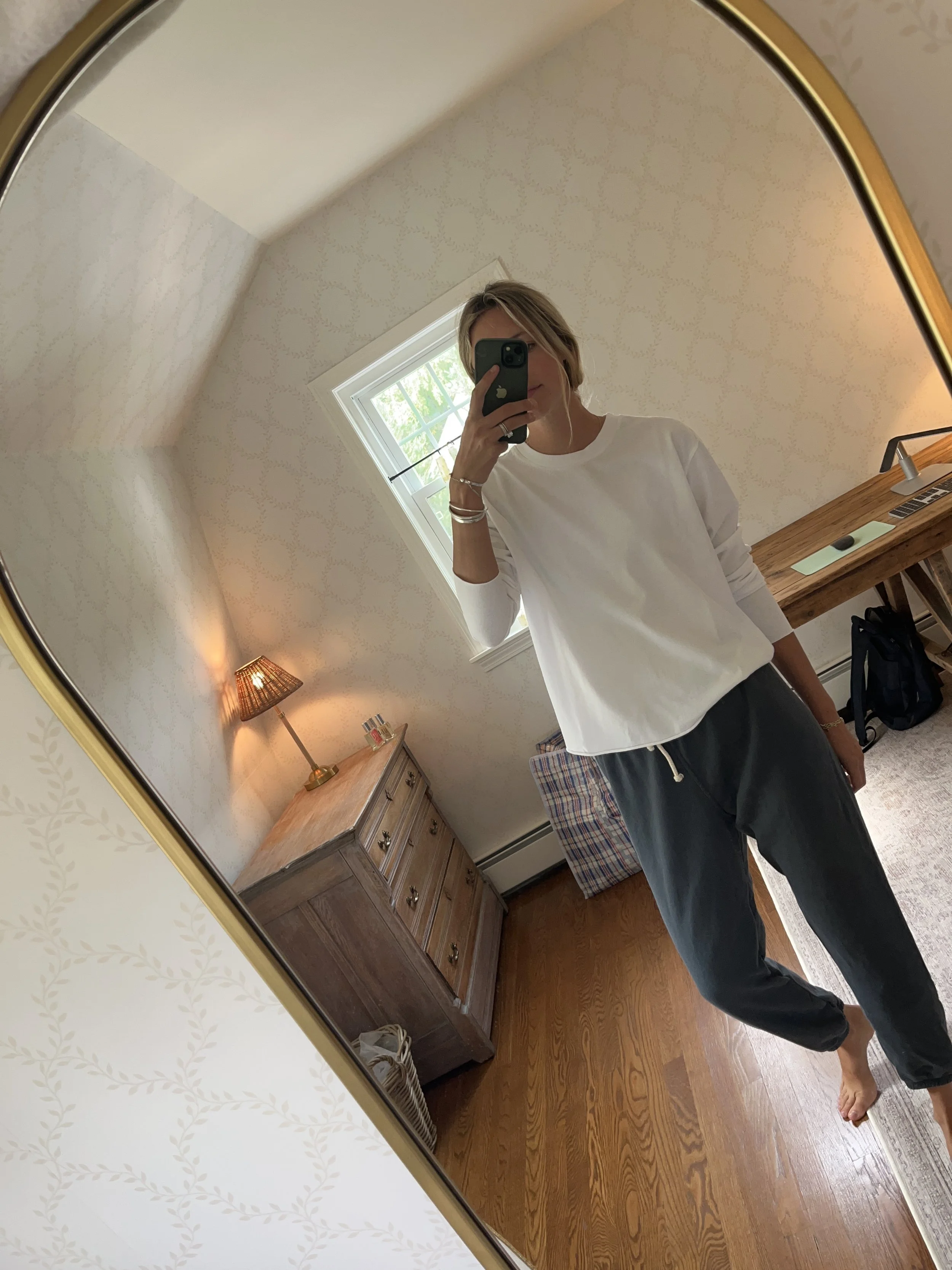 Person taking a mirror selfie in a living room with hardwood floors, wearing a black fleece jacket, gray shirt, black leggings, gray sneakers, and a white cap with a logo.