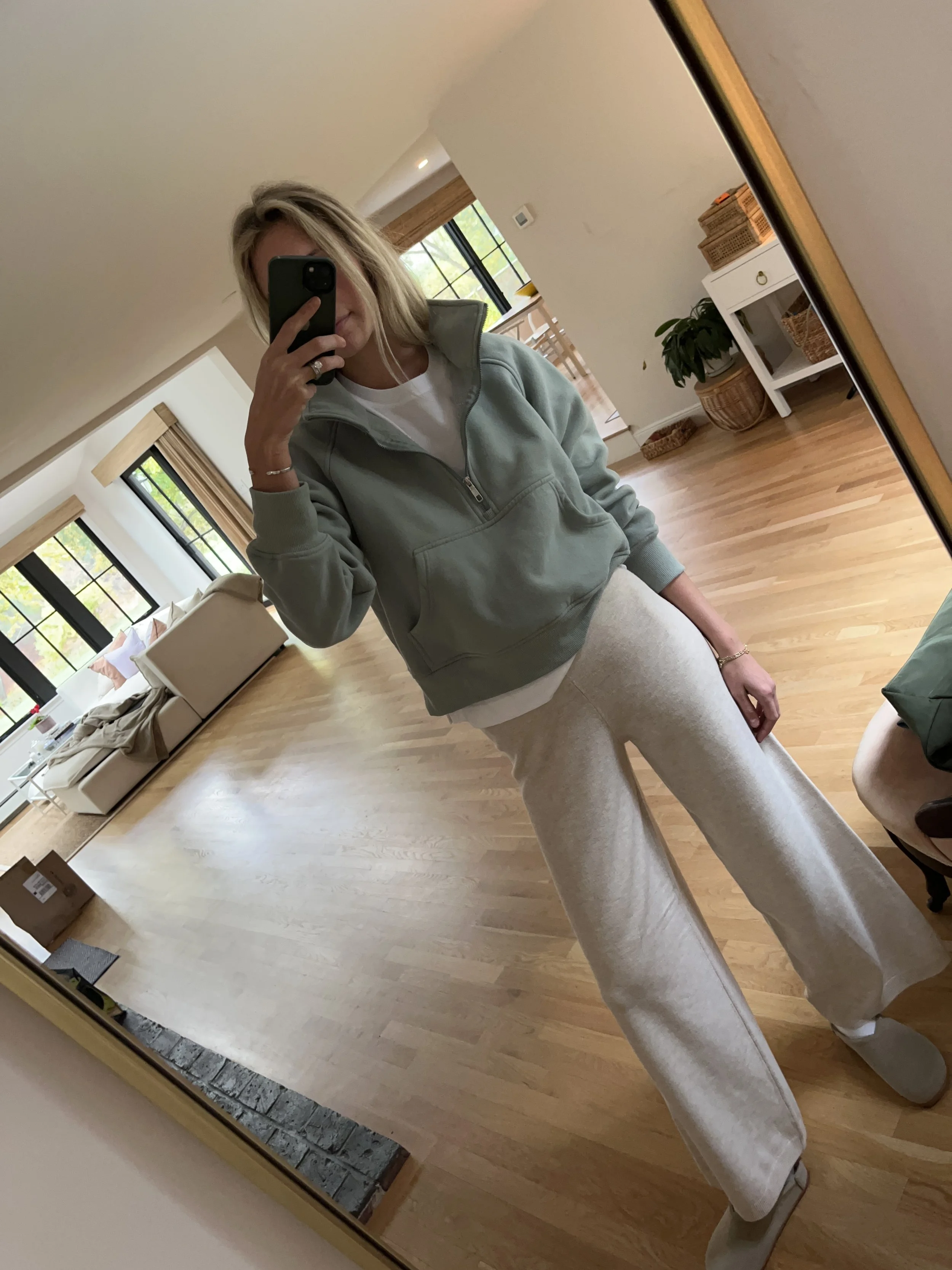 Woman taking a mirror selfie in a spacious living room with wooden floors, large windows, and modern furniture. She is wearing a light gray sweatshirt and beige wide-leg pants.