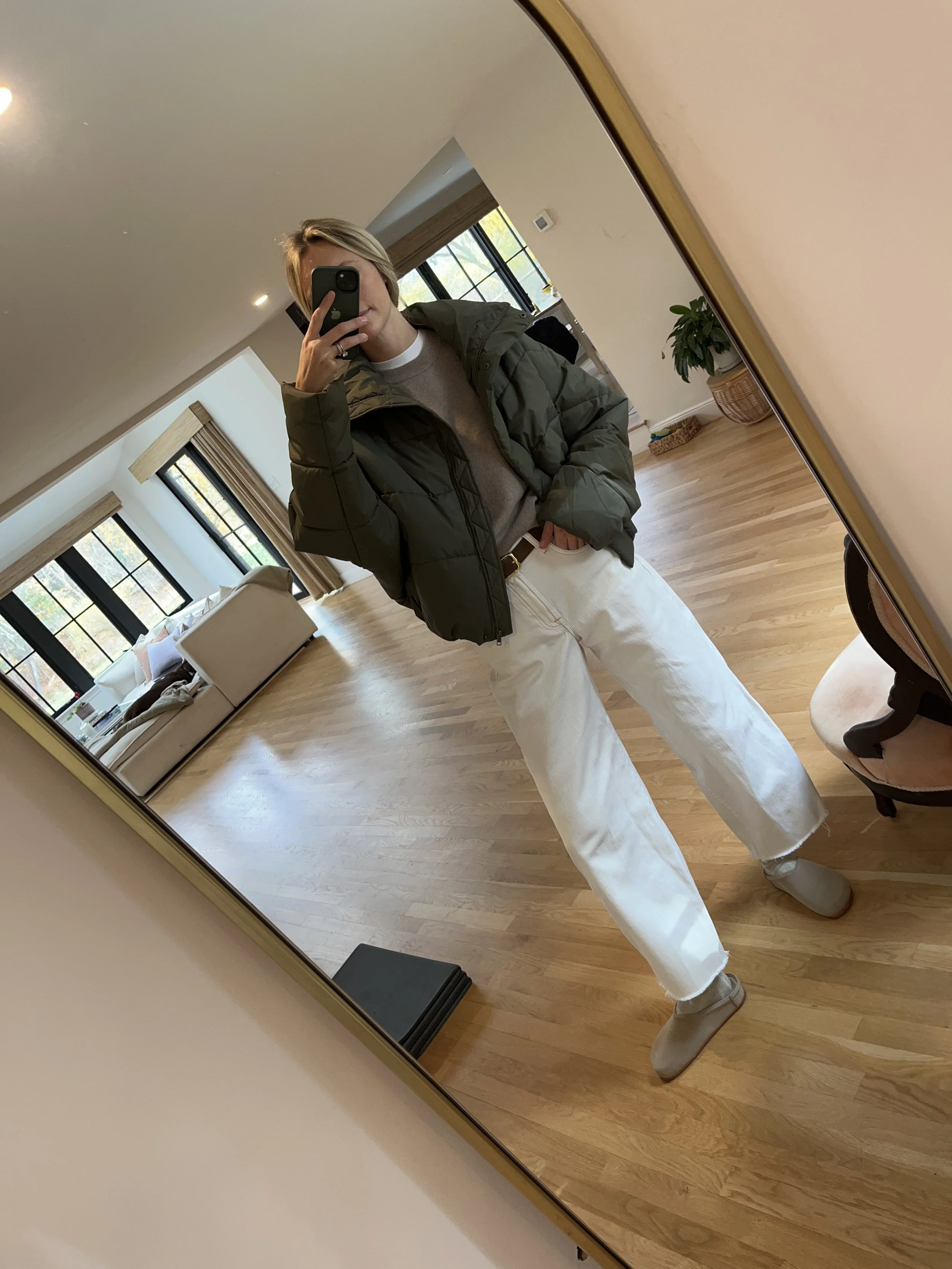 A woman taking a mirror selfie in a spacious, well-lit living room with hardwood floors, large windows, a white sofa, and a side table with a plant.