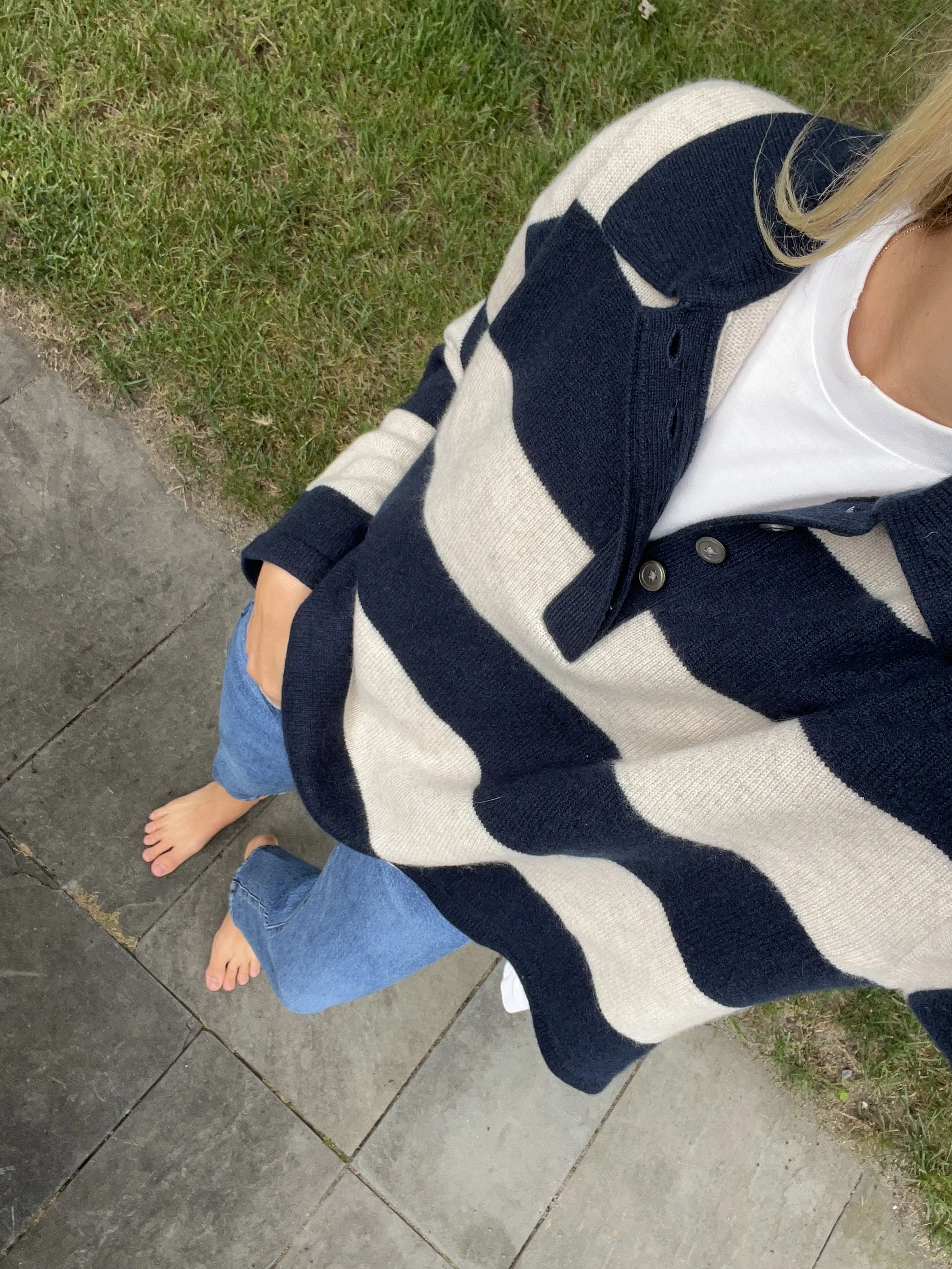 A woman taking a mirror selfie in a spacious living room with wooden floors, large windows, and minimal furniture, wearing a beige jacket, black top, blue jeans, and tan shoes.