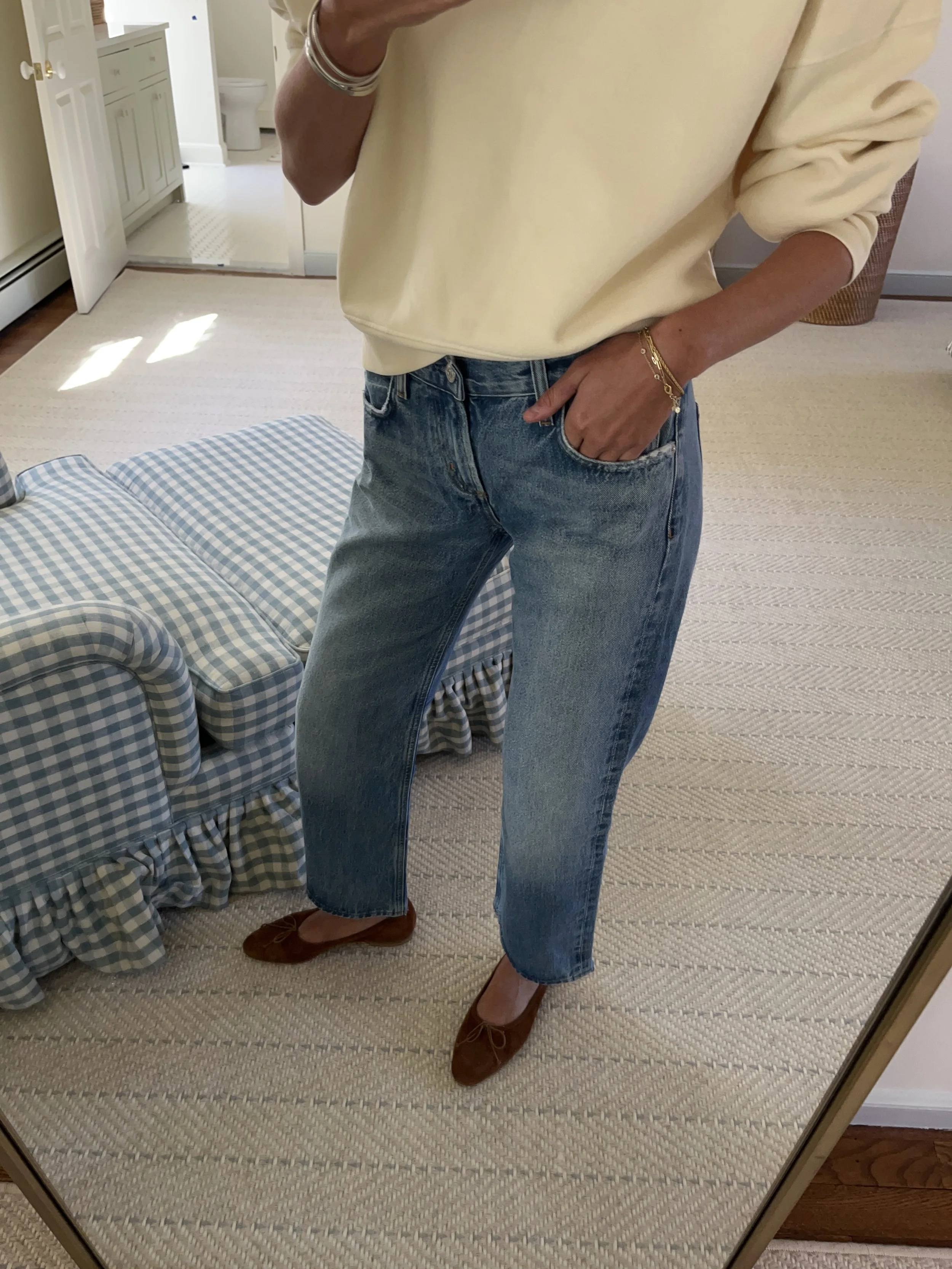 Person taking a mirror selfie in a room with wooden floors, wearing a striped brown and white long-sleeve shirt, light blue jeans, and black and white sneakers.