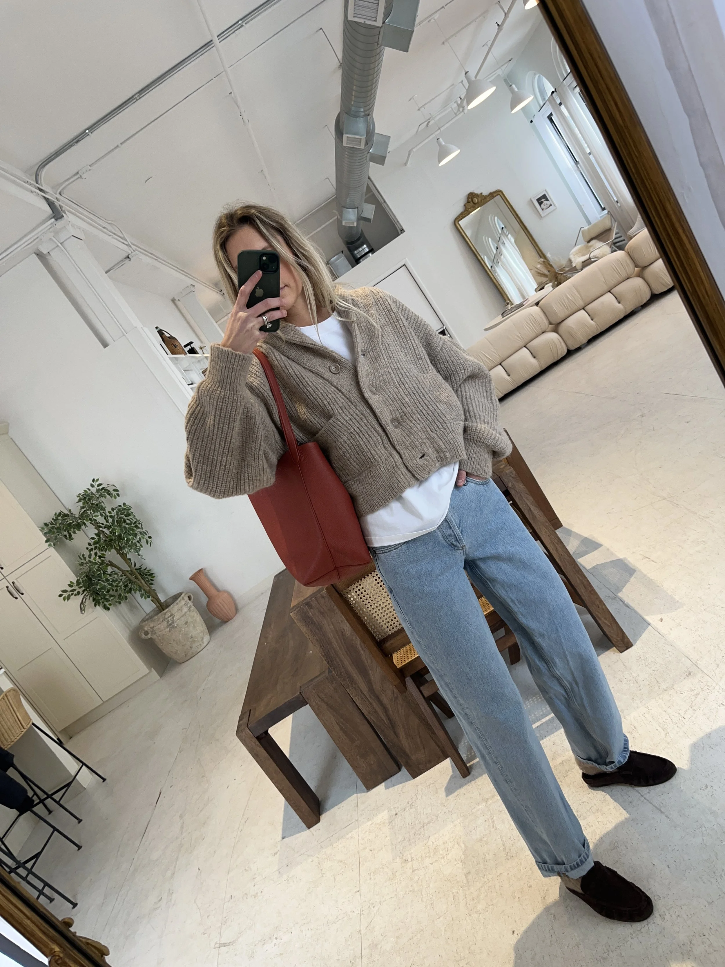 Person taking a mirror selfie in a living room with hardwood floors, wearing a black fleece jacket, gray shirt, black leggings, gray sneakers, and a white cap with a logo.