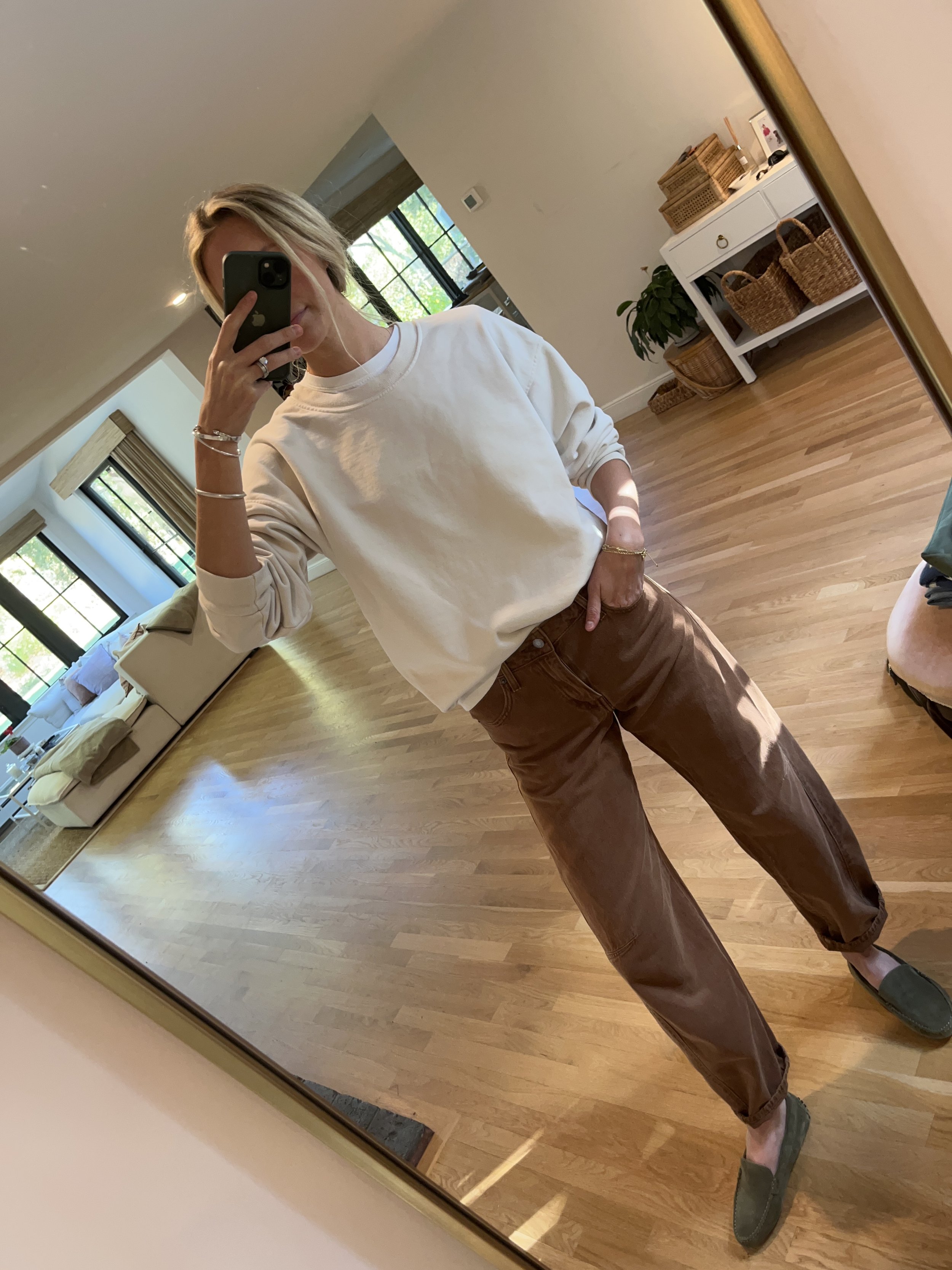 A woman taking a mirror selfie in a bright living room with hardwood floors, large windows, a white and beige sofa, and a white side table with baskets and a potted plant.