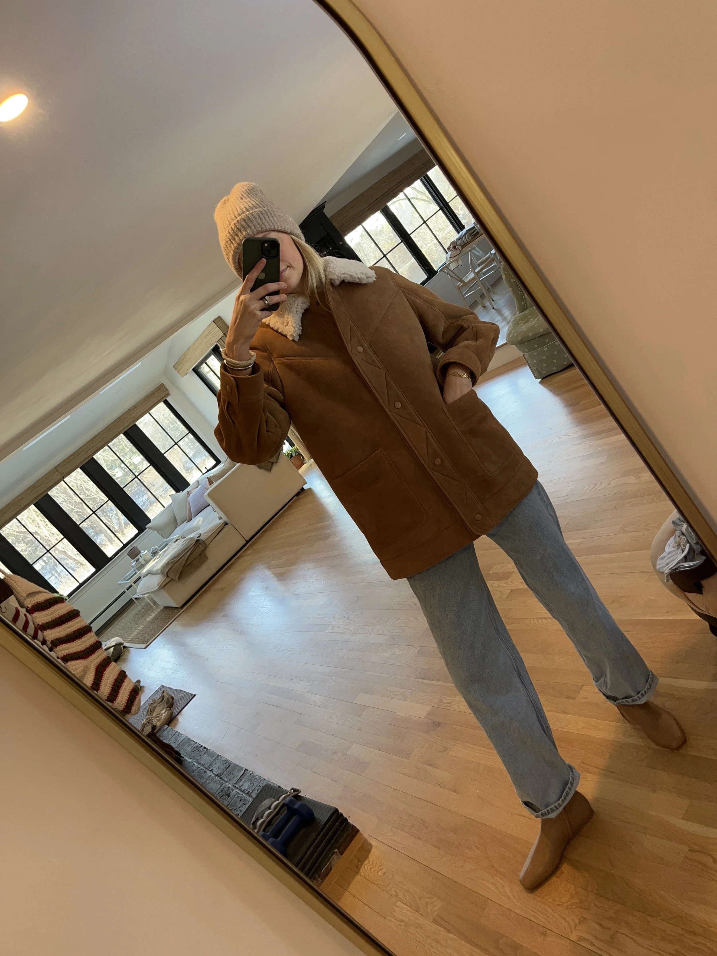 Person taking a mirror selfie in a room with wooden floors, wearing a striped brown and white long-sleeve shirt, light blue jeans, and black and white sneakers.