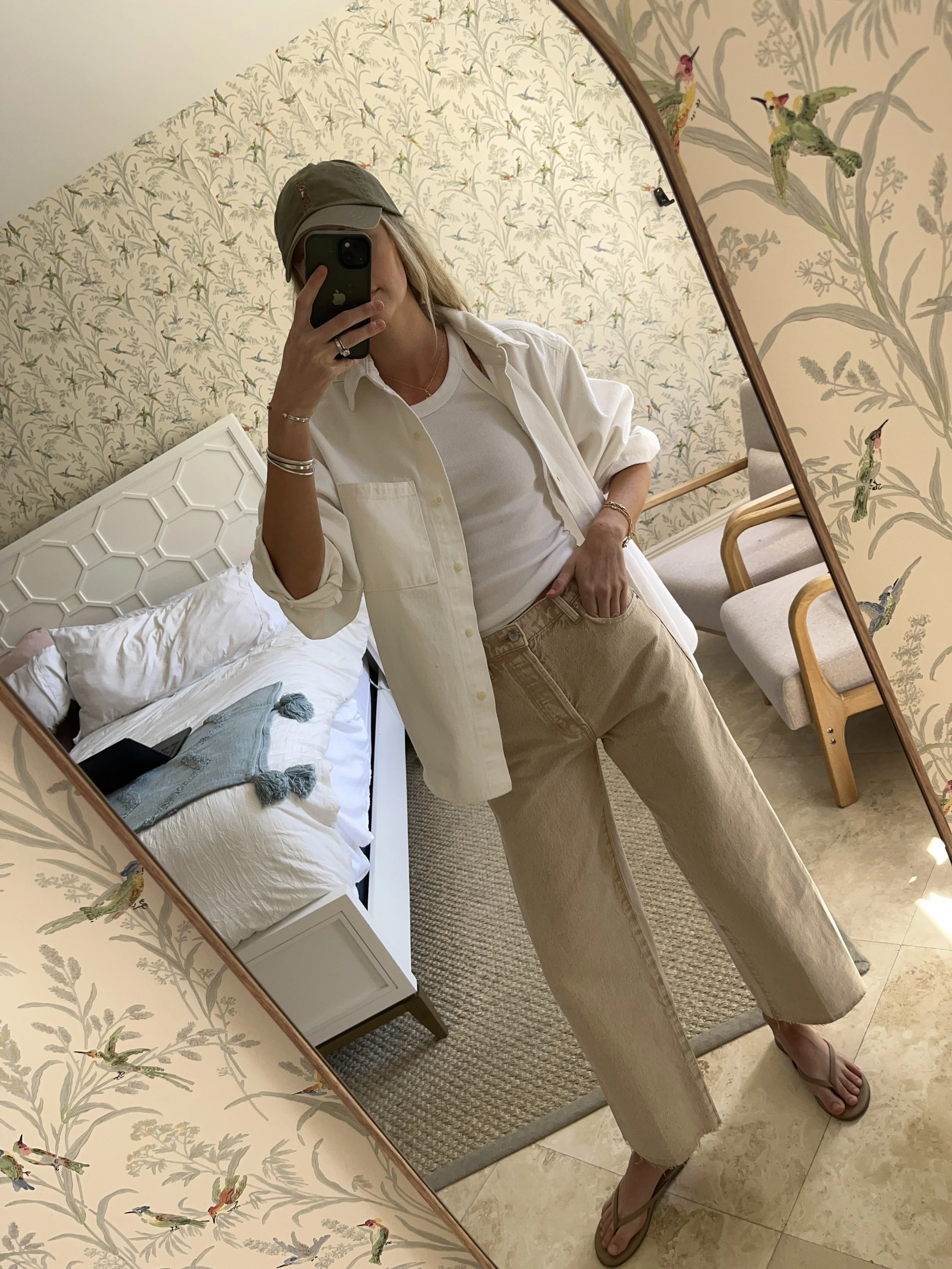 A woman taking a mirror selfie in a spacious, well-lit living room with hardwood floors, large windows, a white sofa, and a side table with a plant.