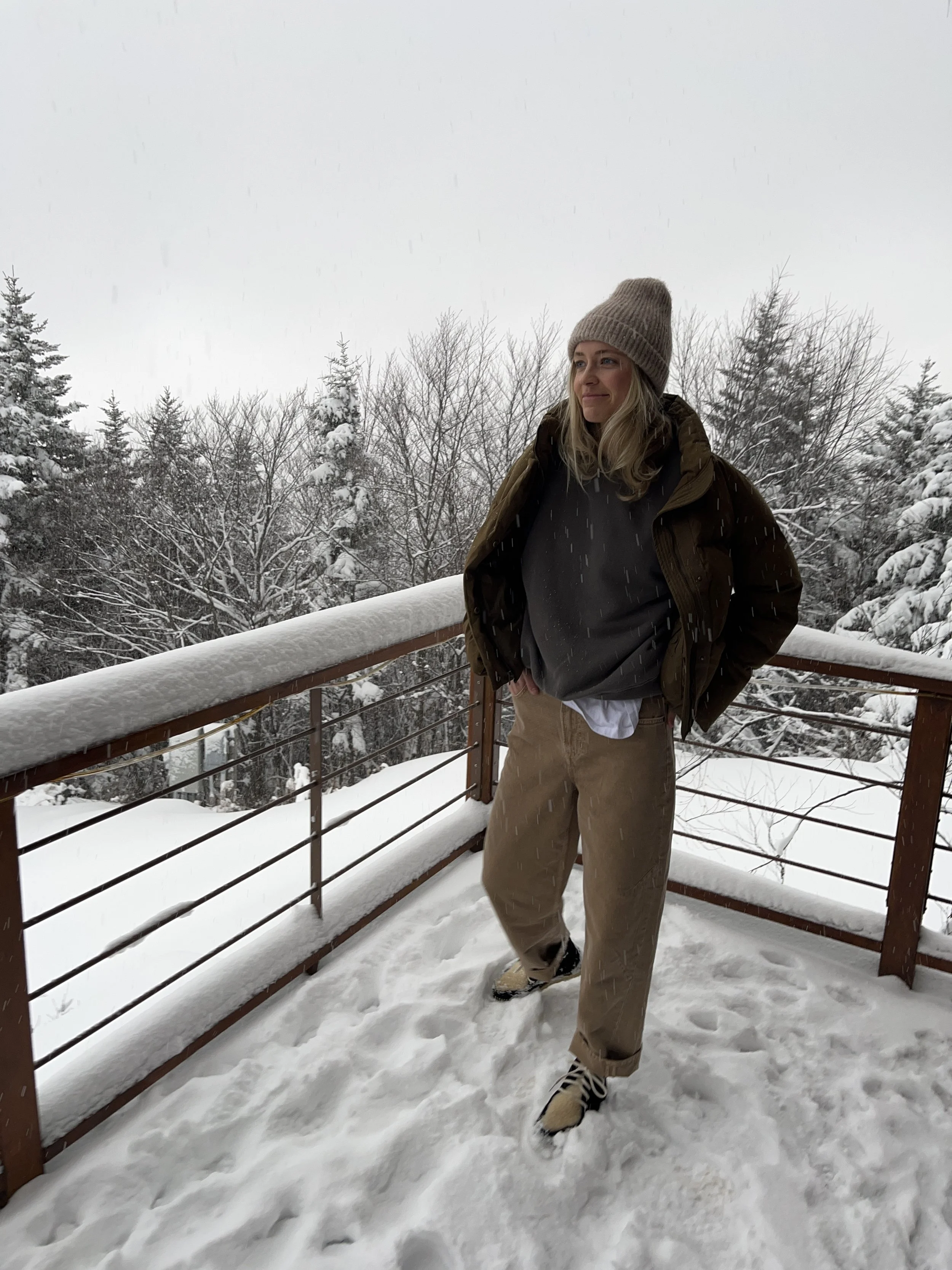 A woman taking a mirror selfie in a spacious living room with wooden floors, large windows, and minimal furniture, wearing a beige jacket, black top, blue jeans, and tan shoes.