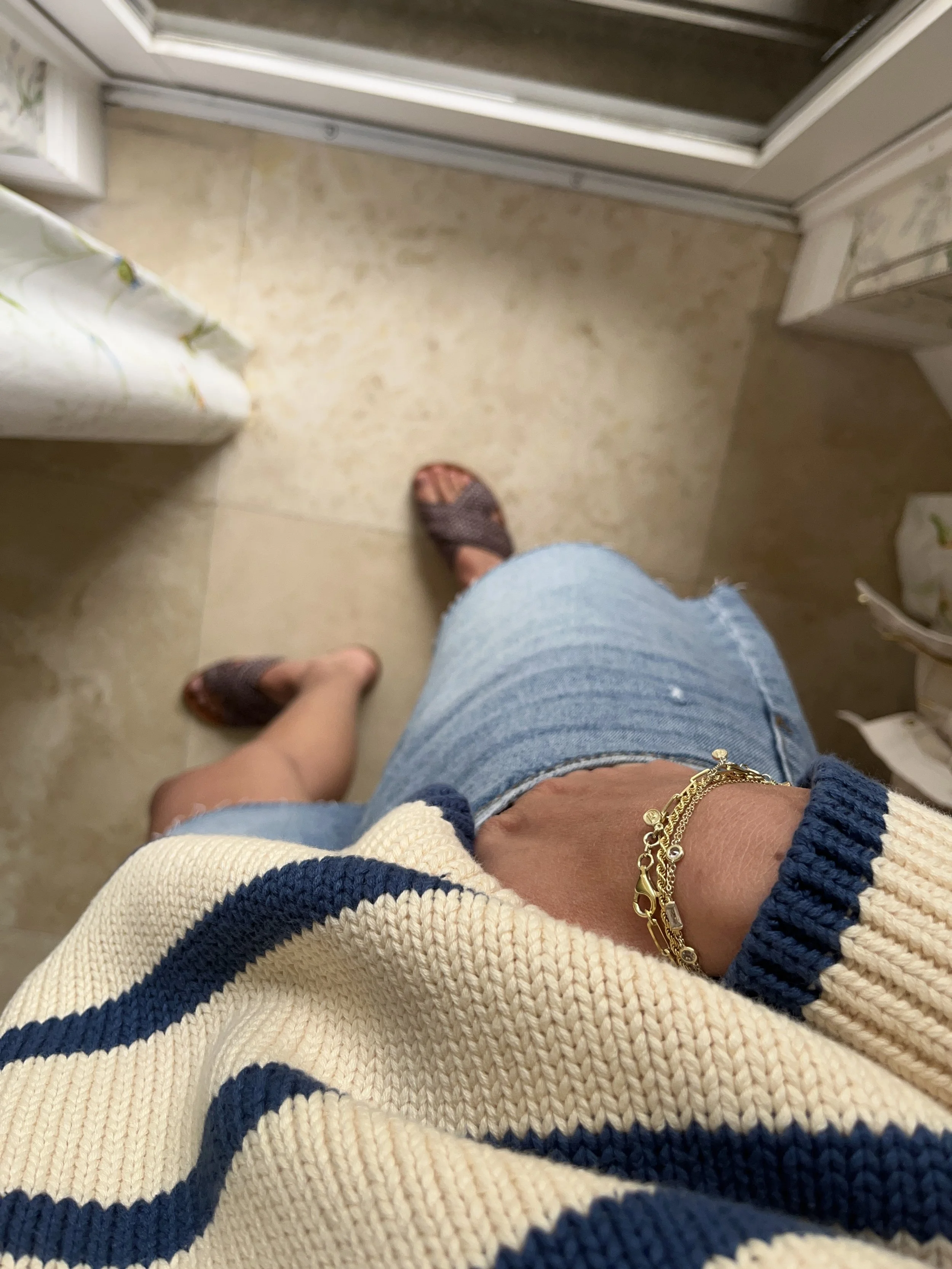 A woman taking a mirror selfie in a spacious living room with wooden floors, large windows, and minimal furniture, wearing a beige jacket, black top, blue jeans, and tan shoes.