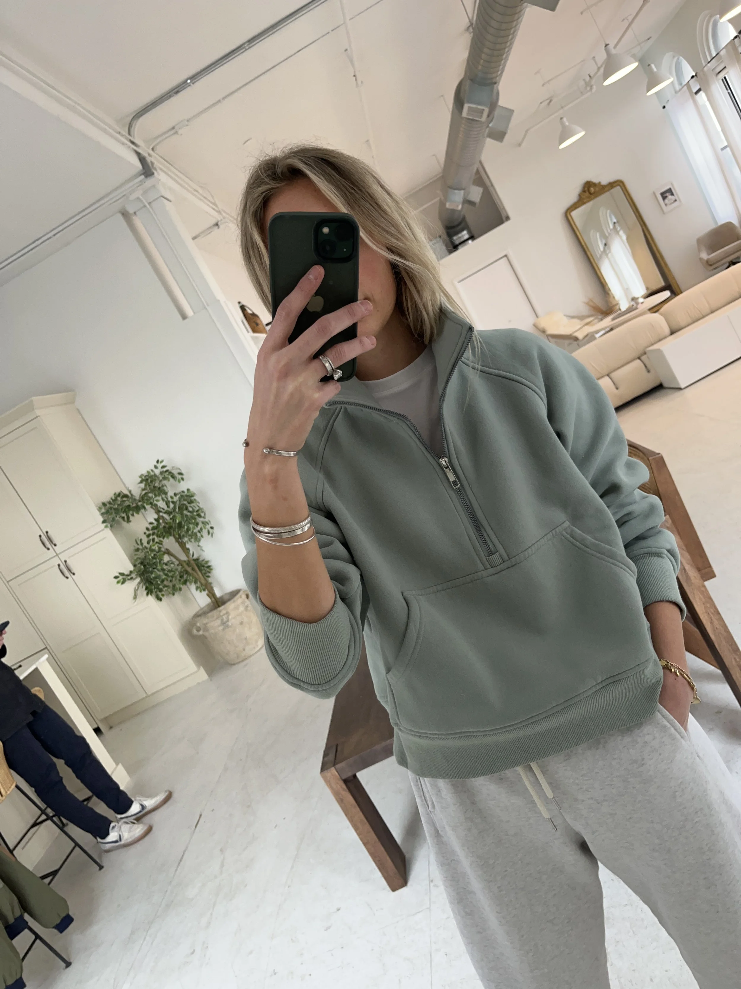 A woman taking a mirror selfie in a spacious, well-lit living room with hardwood floors, large windows, a white sofa, and a side table with a plant.