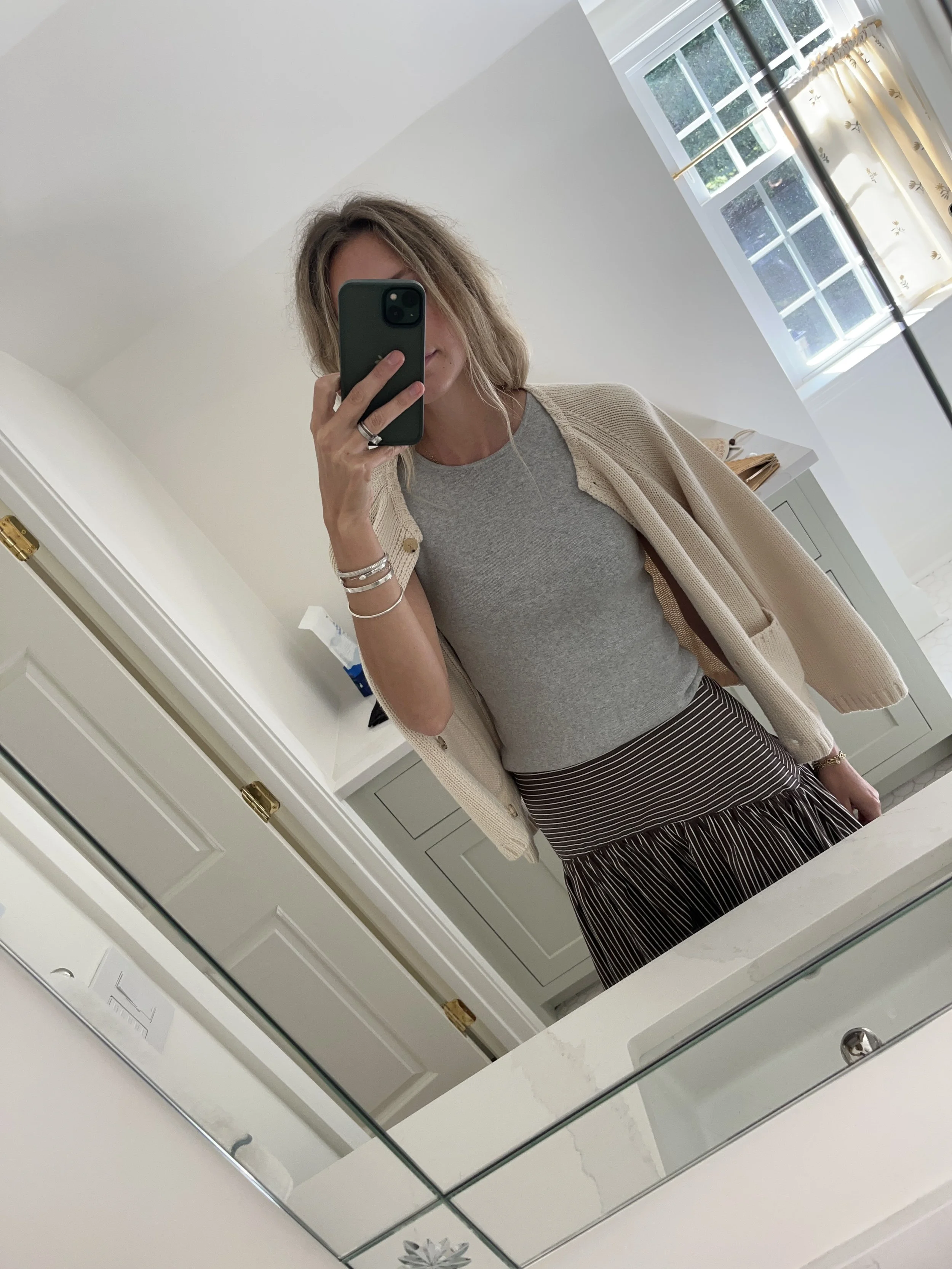 Person taking a mirror selfie in a living room with hardwood floors, wearing a black fleece jacket, gray shirt, black leggings, gray sneakers, and a white cap with a logo.