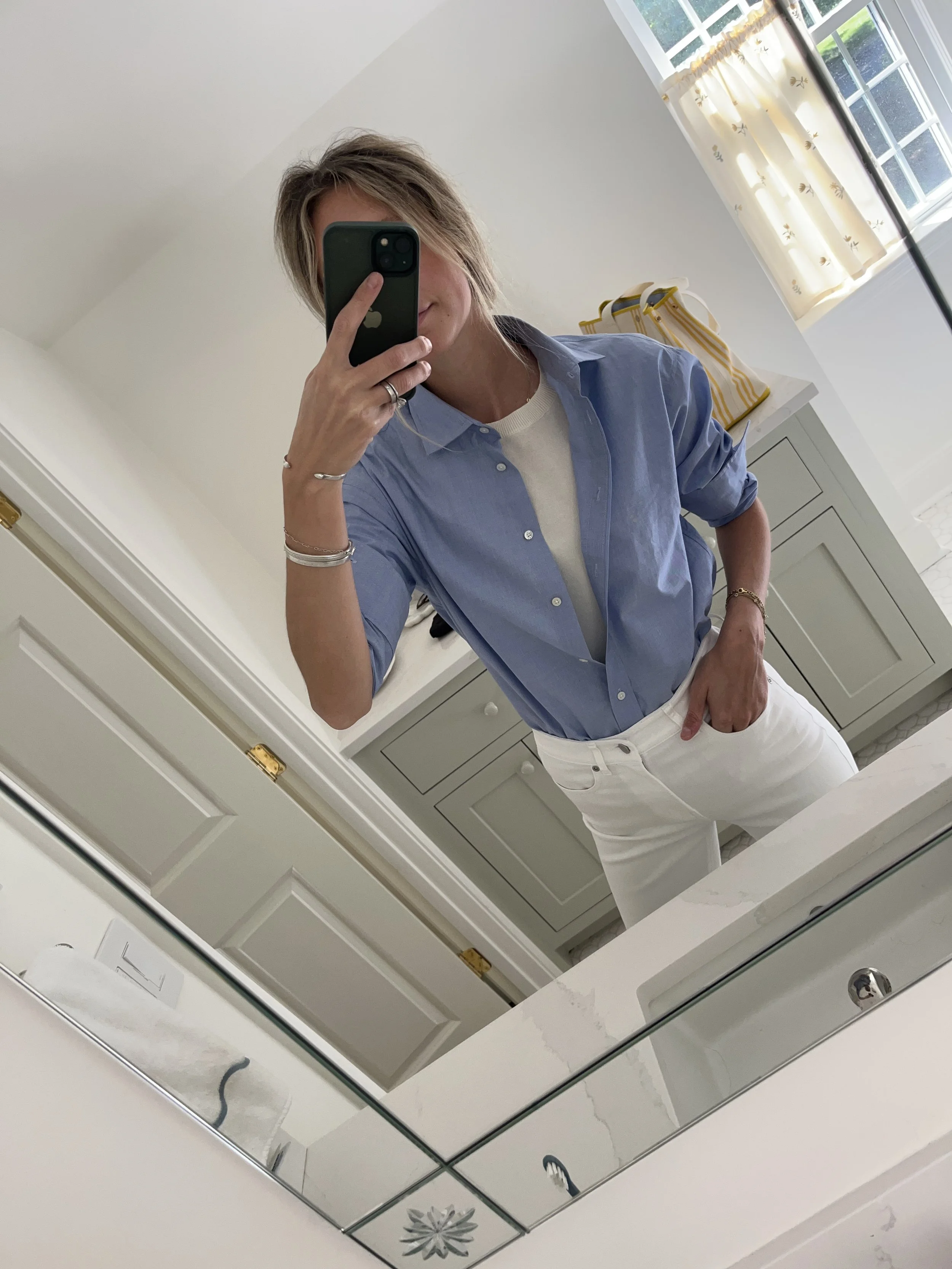 A woman taking a mirror selfie in a spacious, well-lit living room with hardwood floors, large windows, a white sofa, and a side table with a plant.