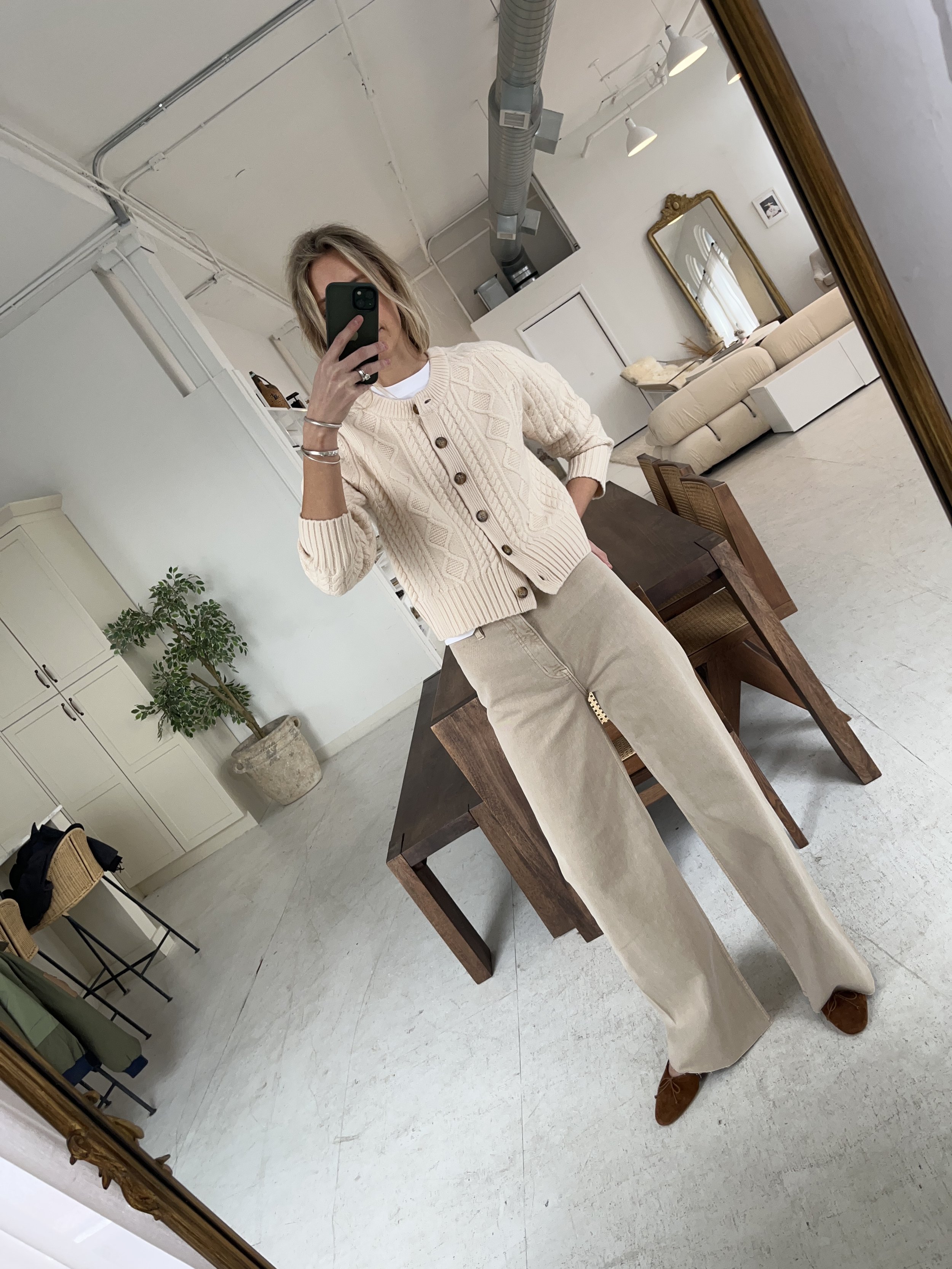A woman taking a mirror selfie in a spacious, well-lit living room with hardwood floors, large windows, a white sofa, and a side table with a plant.