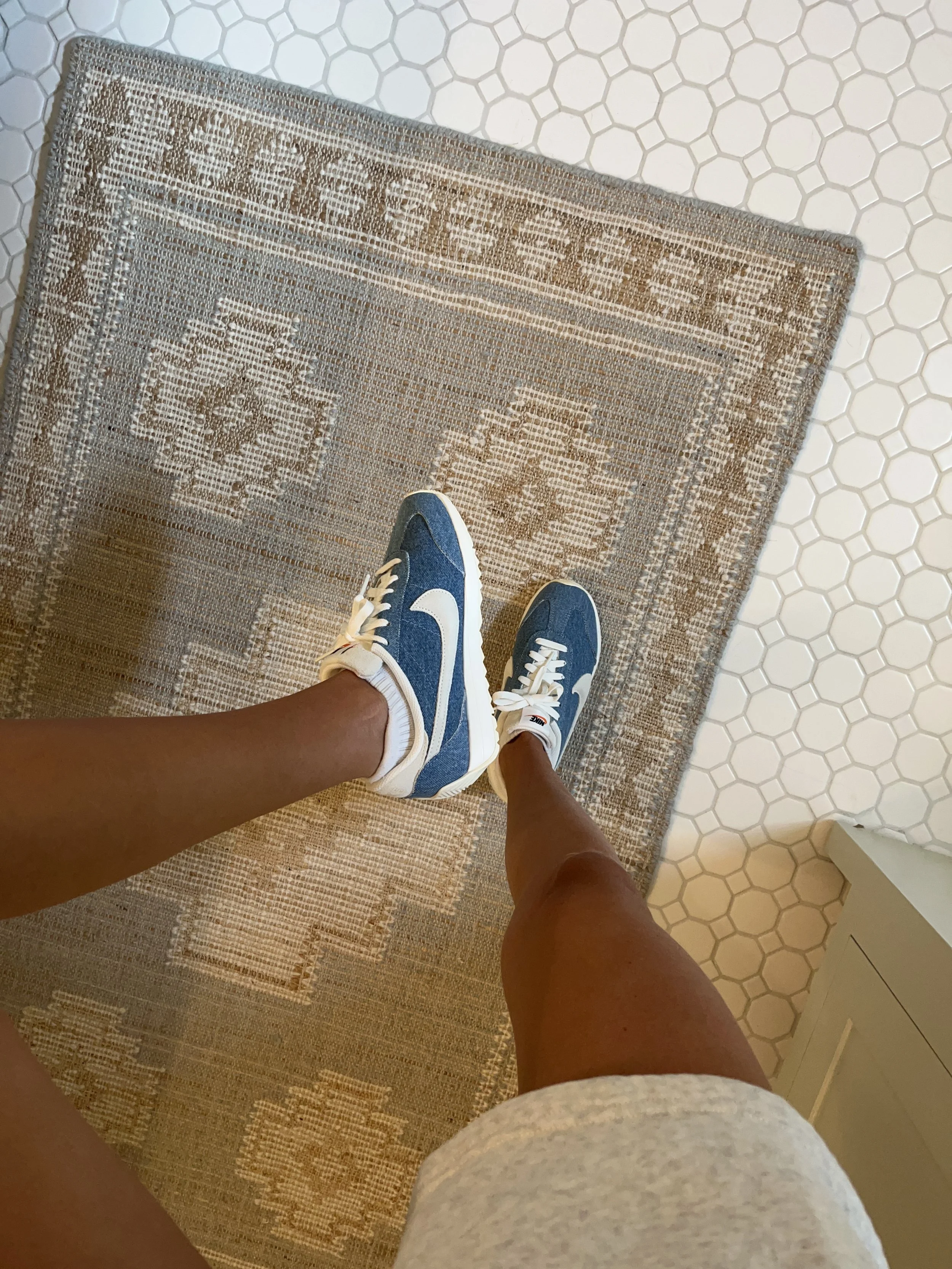 Person taking a mirror selfie in a living room with hardwood floors, wearing a black fleece jacket, gray shirt, black leggings, gray sneakers, and a white cap with a logo.