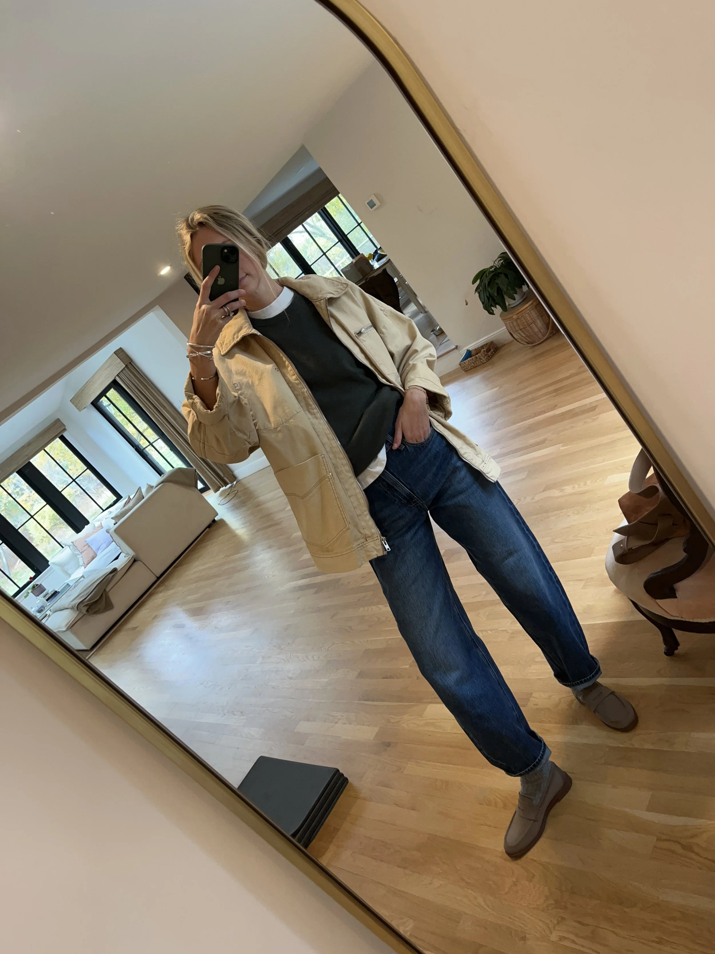A woman taking a mirror selfie in a spacious living room with wooden floors, large windows, and minimal furniture, wearing a beige jacket, black top, blue jeans, and tan shoes.