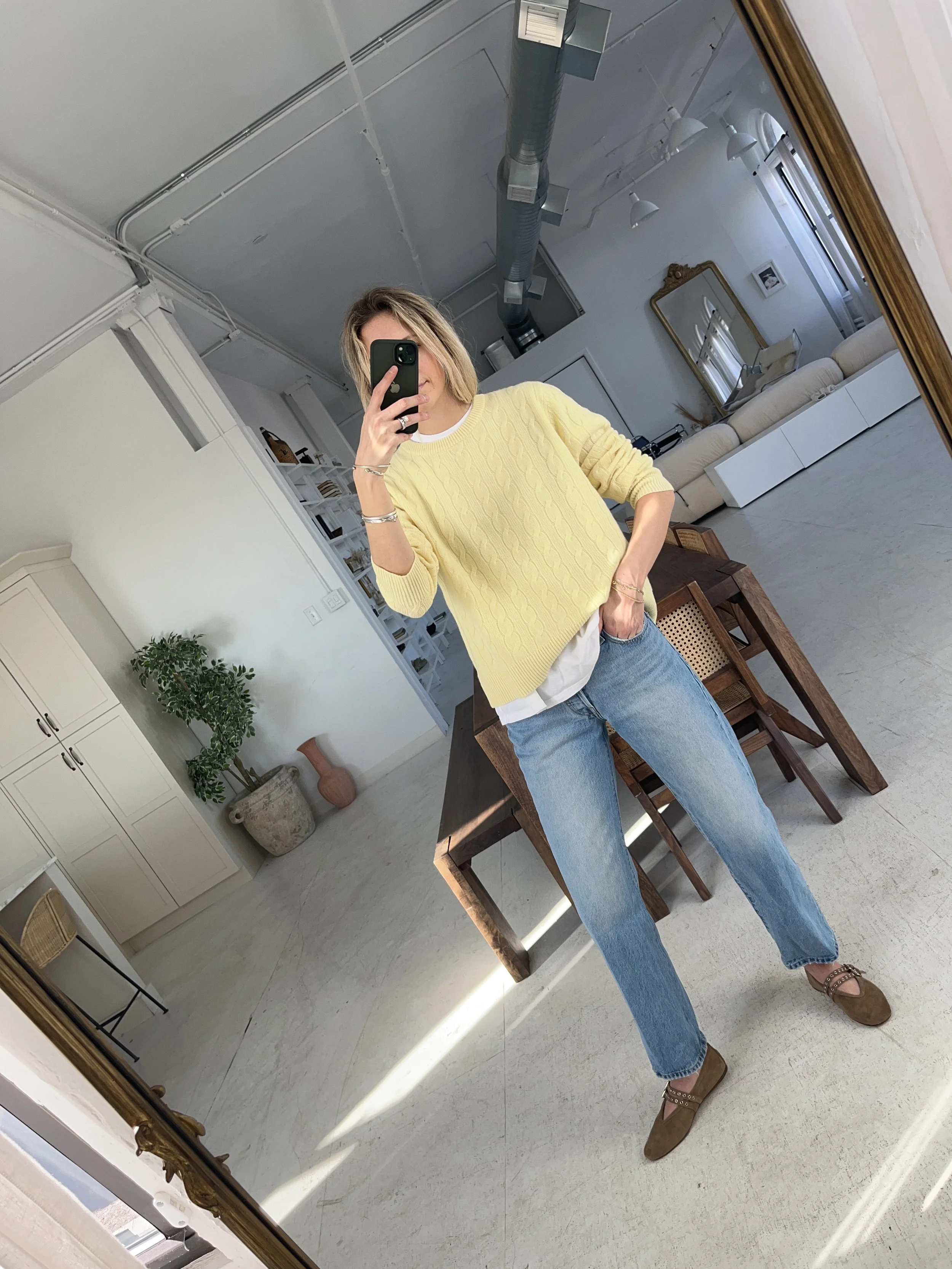 A woman taking a mirror selfie in a spacious, well-lit living room with hardwood floors, large windows, a white sofa, and a side table with a plant.