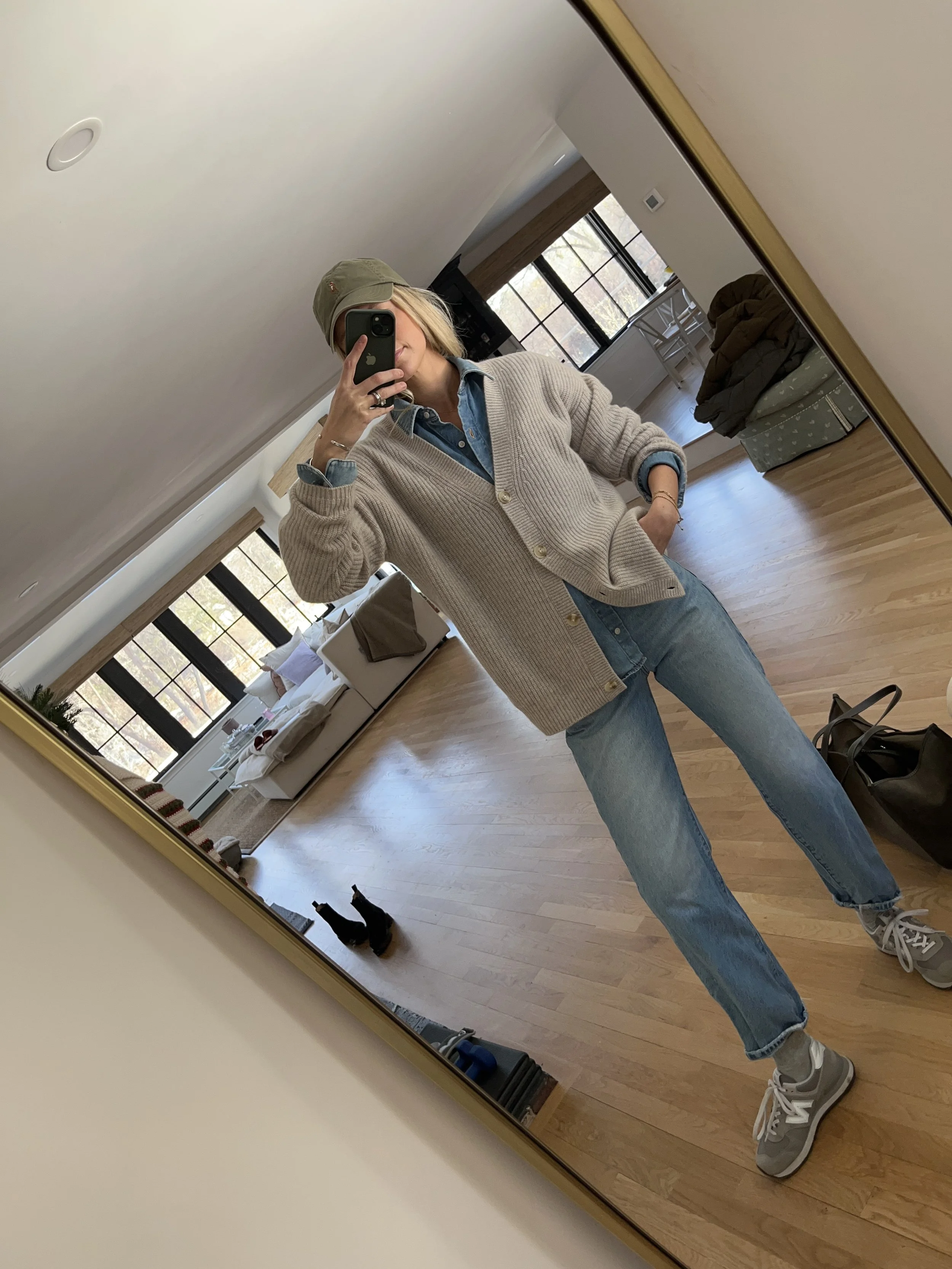 A woman taking a mirror selfie in a spacious, well-lit living room with hardwood floors, large windows, a white sofa, and a side table with a plant.