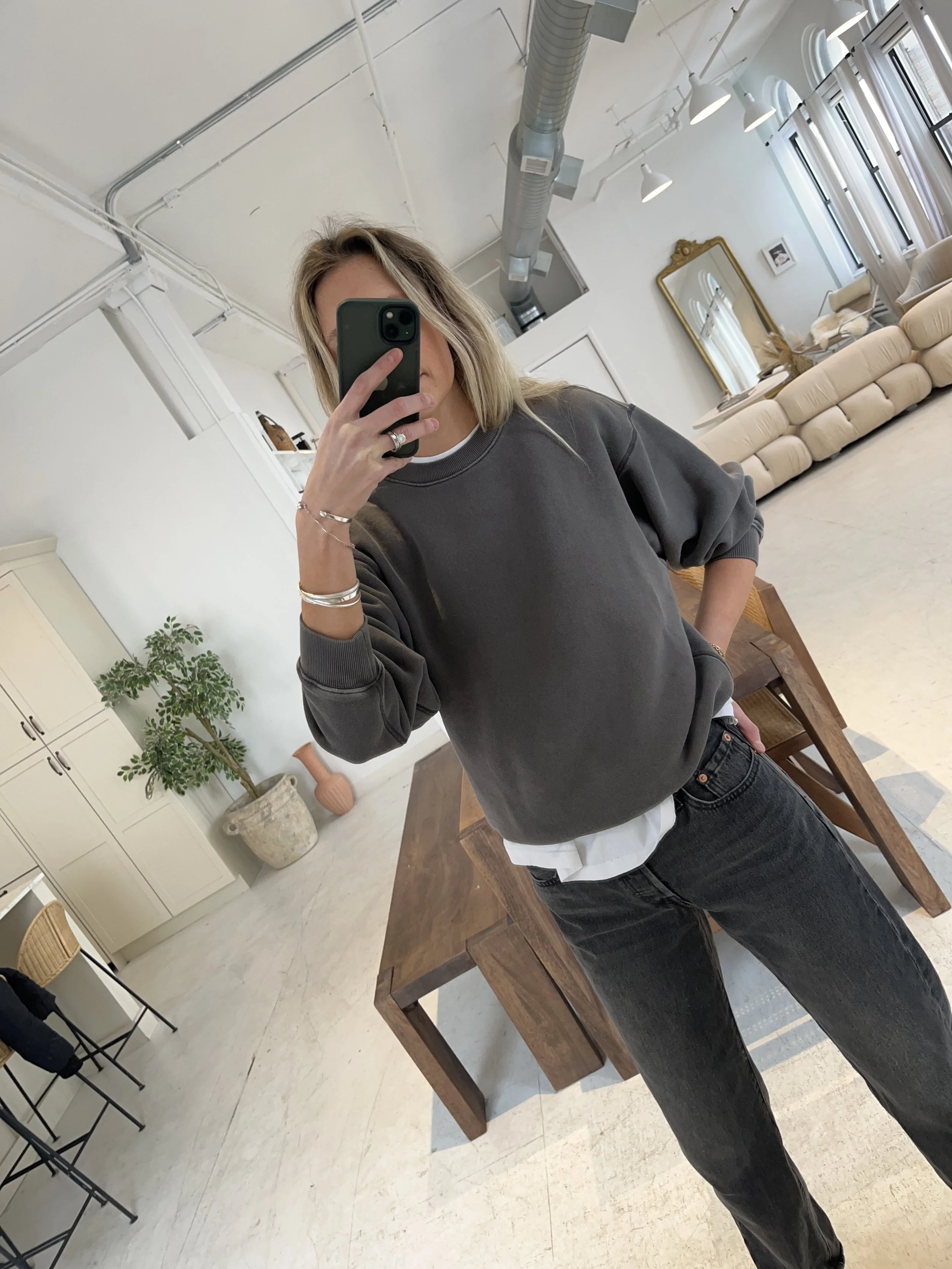 A woman taking a mirror selfie in a spacious living room with wooden floors, large windows, and minimal furniture, wearing a beige jacket, black top, blue jeans, and tan shoes.