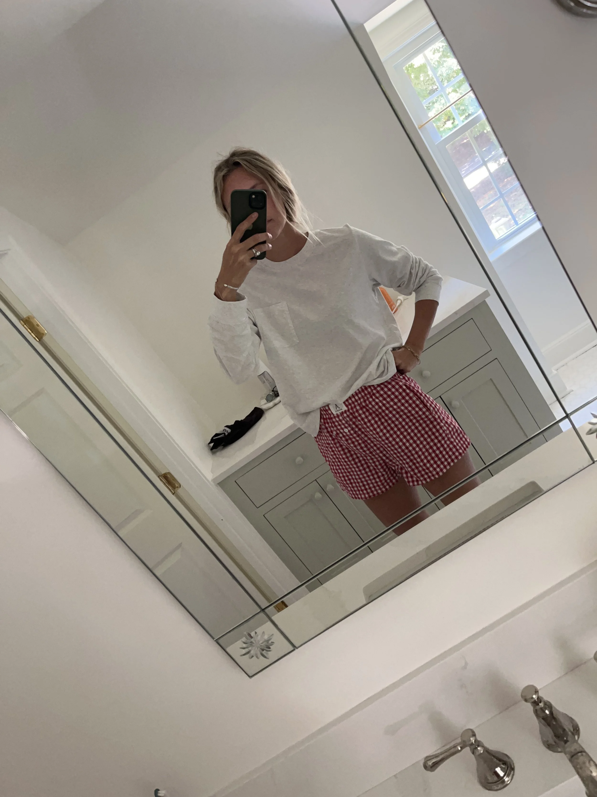 Person taking a mirror selfie in a living room with hardwood floors, wearing a black fleece jacket, gray shirt, black leggings, gray sneakers, and a white cap with a logo.