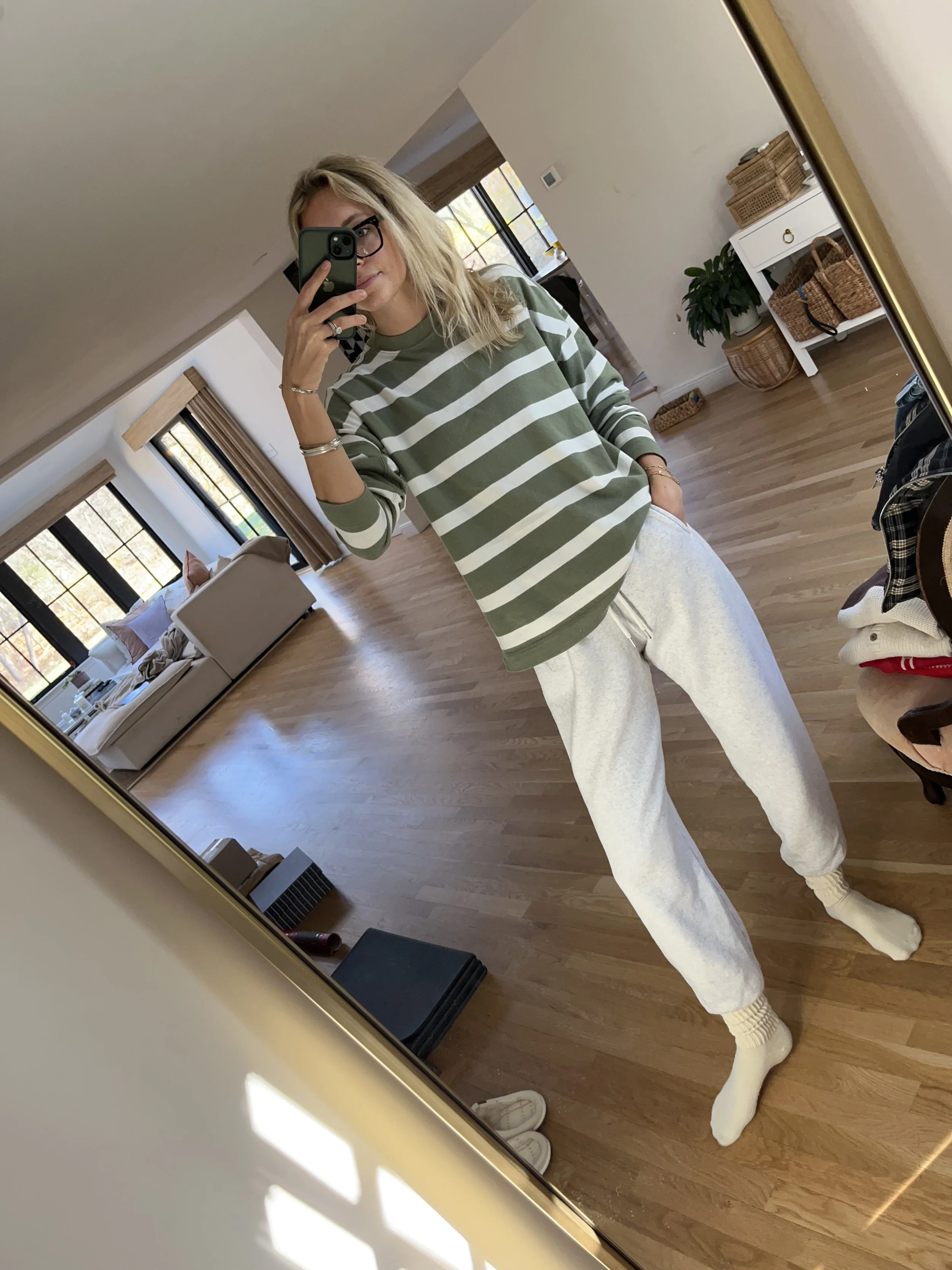Person taking a mirror selfie in a room with wooden floors, wearing a striped brown and white long-sleeve shirt, light blue jeans, and black and white sneakers.